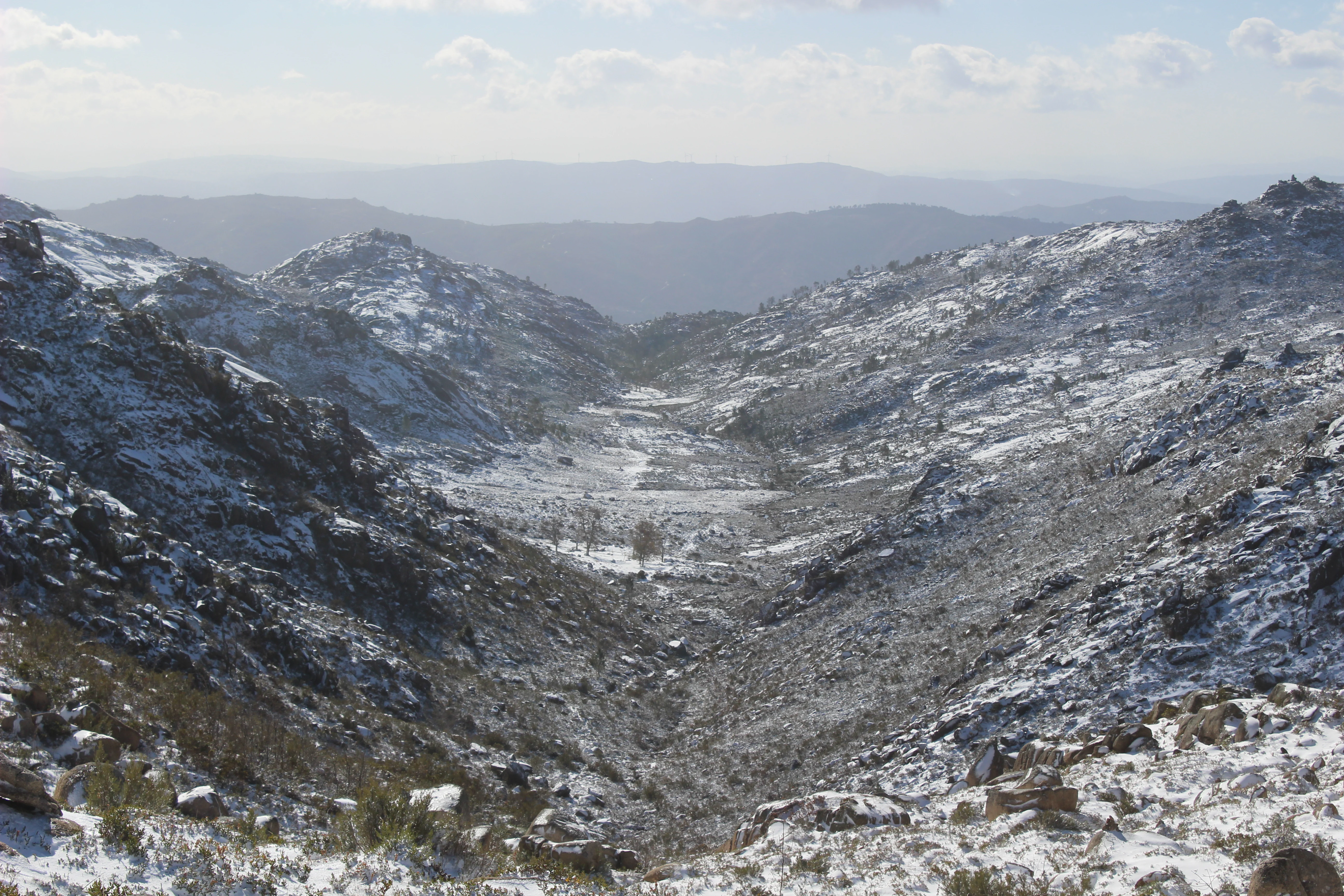 Snow-covered mountain valley with rocky slopes and distant hills under a partly cloudy sky