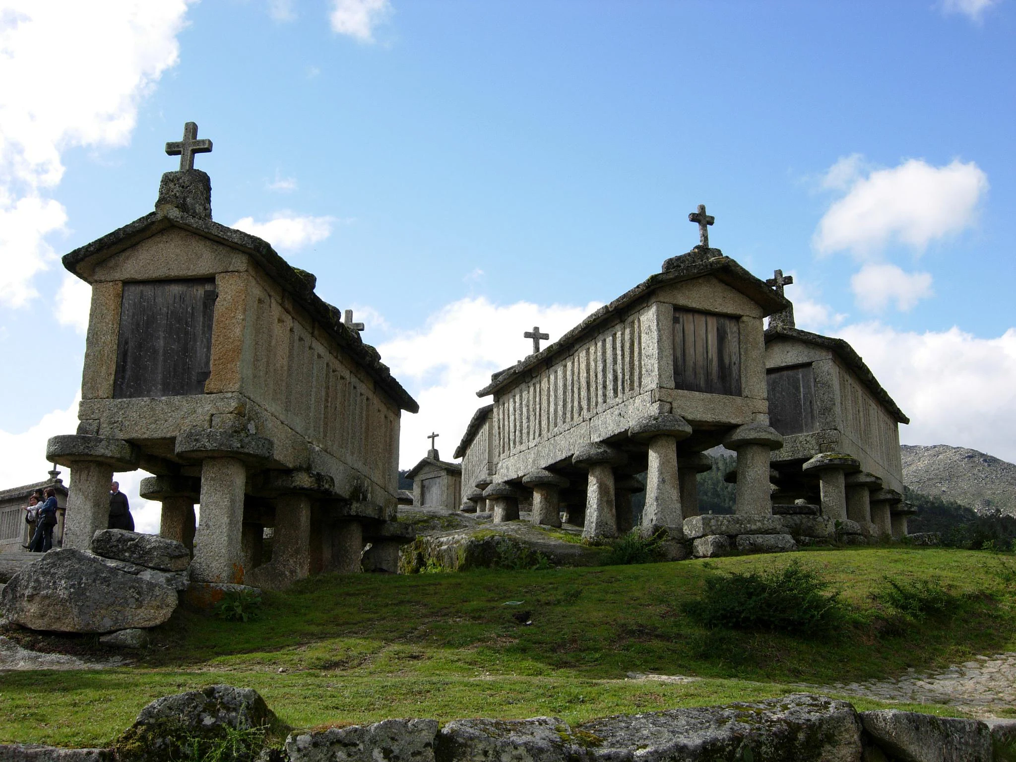 Stone granaries elevated on pillars with crosses on top, set against a mountainous backdrop in a grassy area