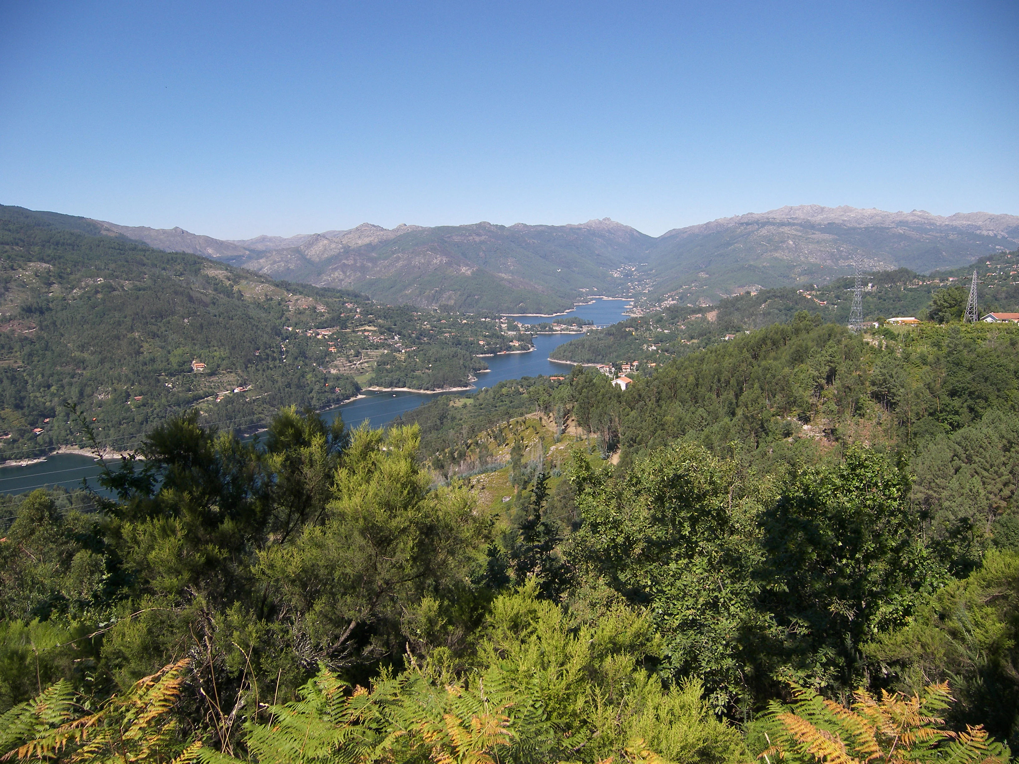 Wide landscape view of a mountain valley with a lake surrounded by forested hills and distant mountains under a clear blue sky