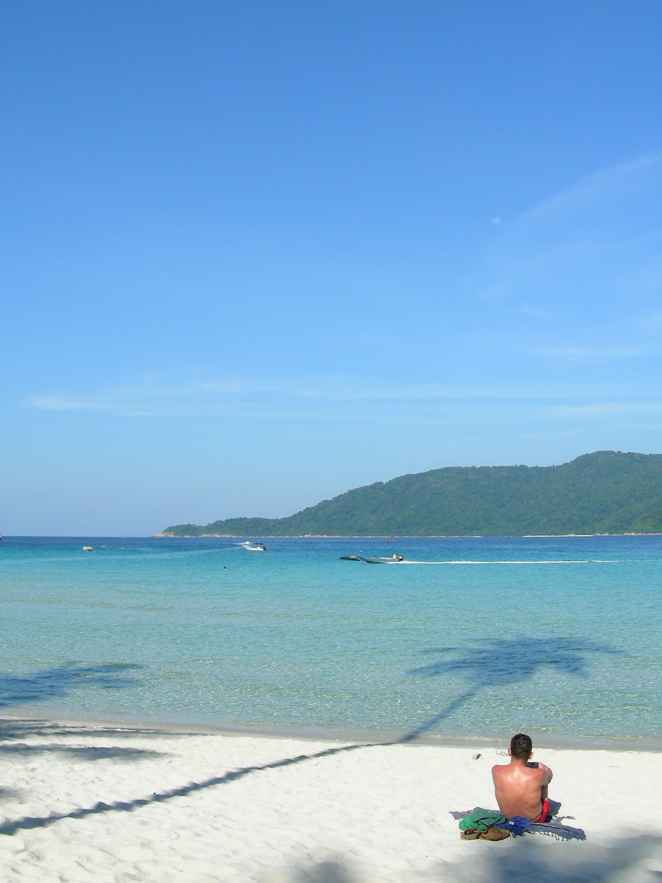 A person sits on a white sand beach facing the turquoise ocean with a distant forested island visible under a clear blue sky; shadows of palm trees are cast across the foreground sand.
