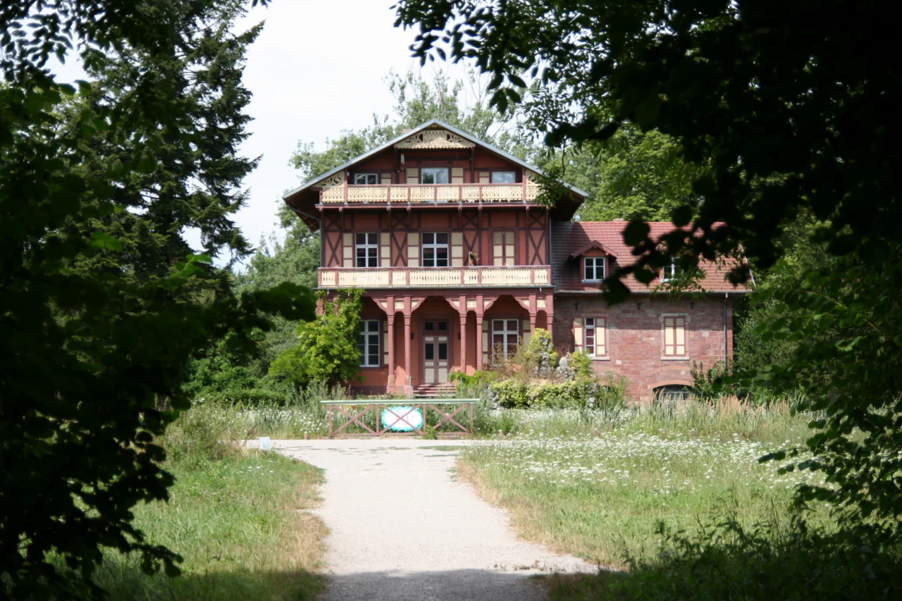 A historic red brick and half-timbered building framed by dark green foliage, featuring a dirt path leading to the entrance.