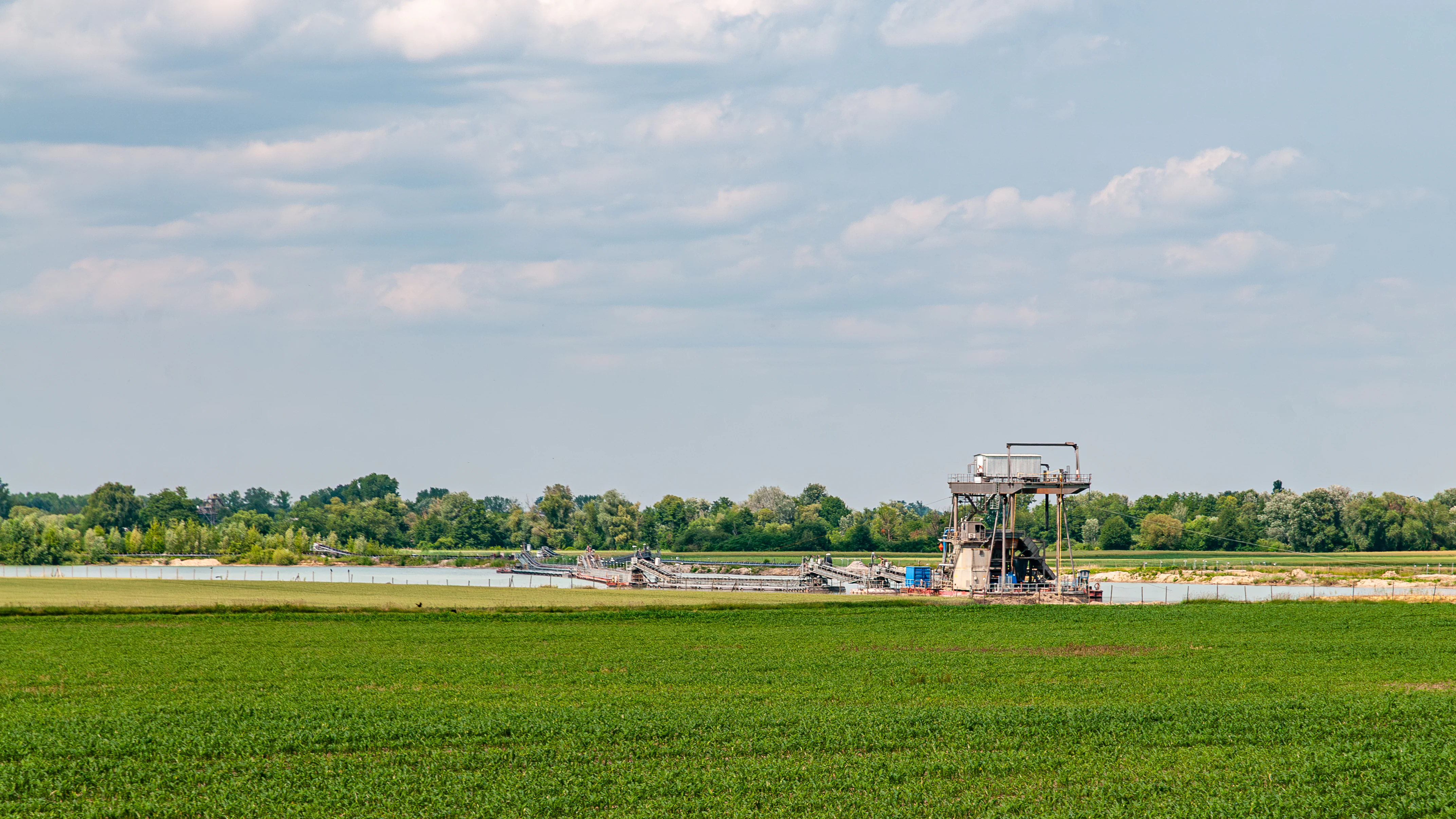 A gravel mining installation with machinery sits on the edge of a water body, surrounded by green fields and trees under a cloudy sky.