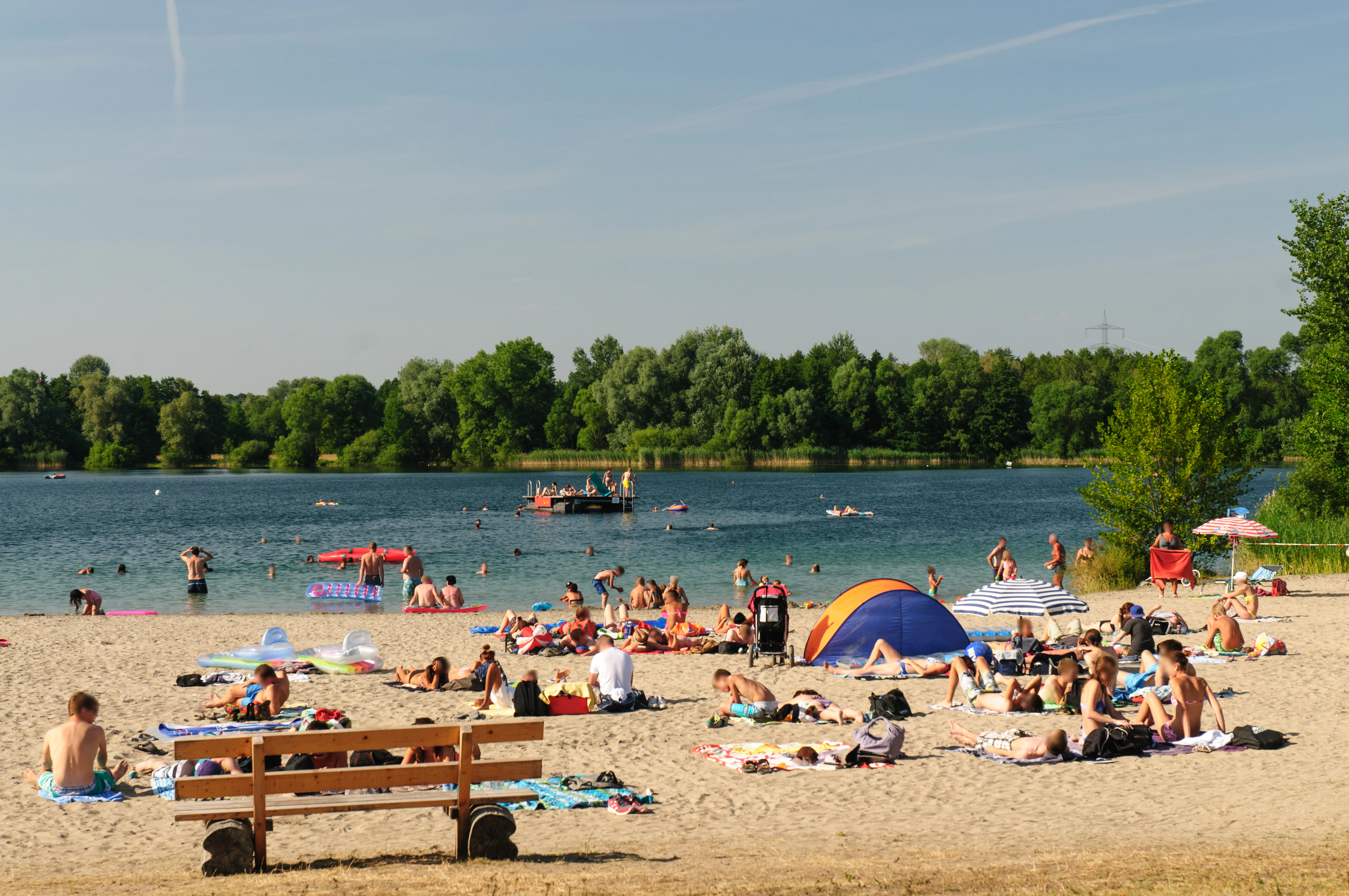 A sandy beach at a lake with many people relaxing, sunbathing, and swimming on a sunny summer day.