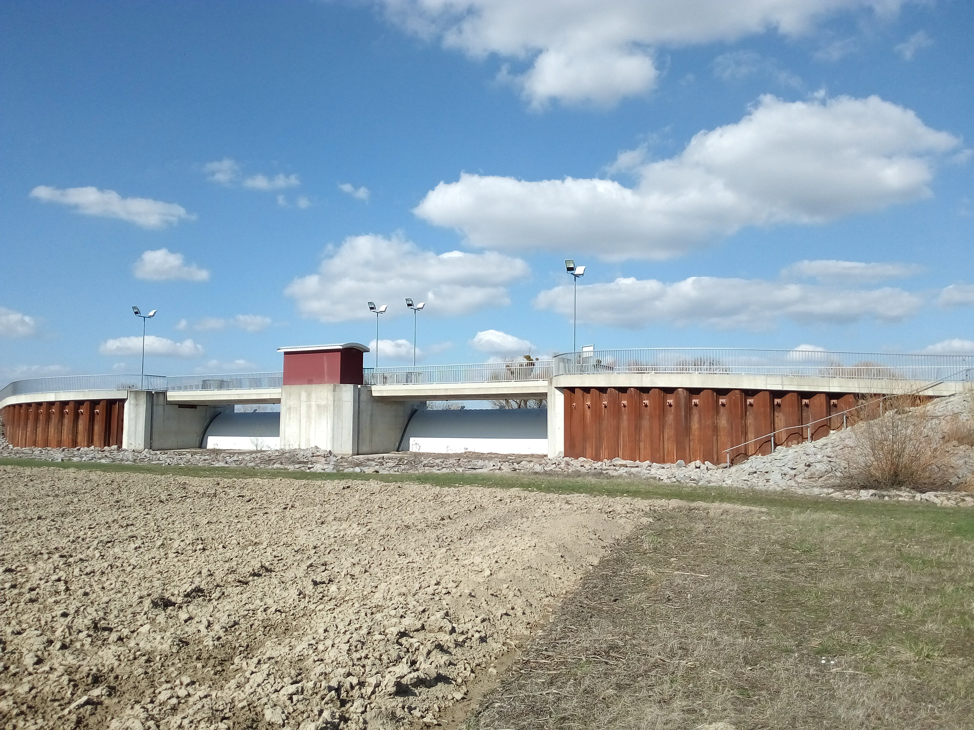 A concrete and steel water management structure stretches horizontally across the scene under a blue sky with scattered clouds, with a tilled field in the foreground.