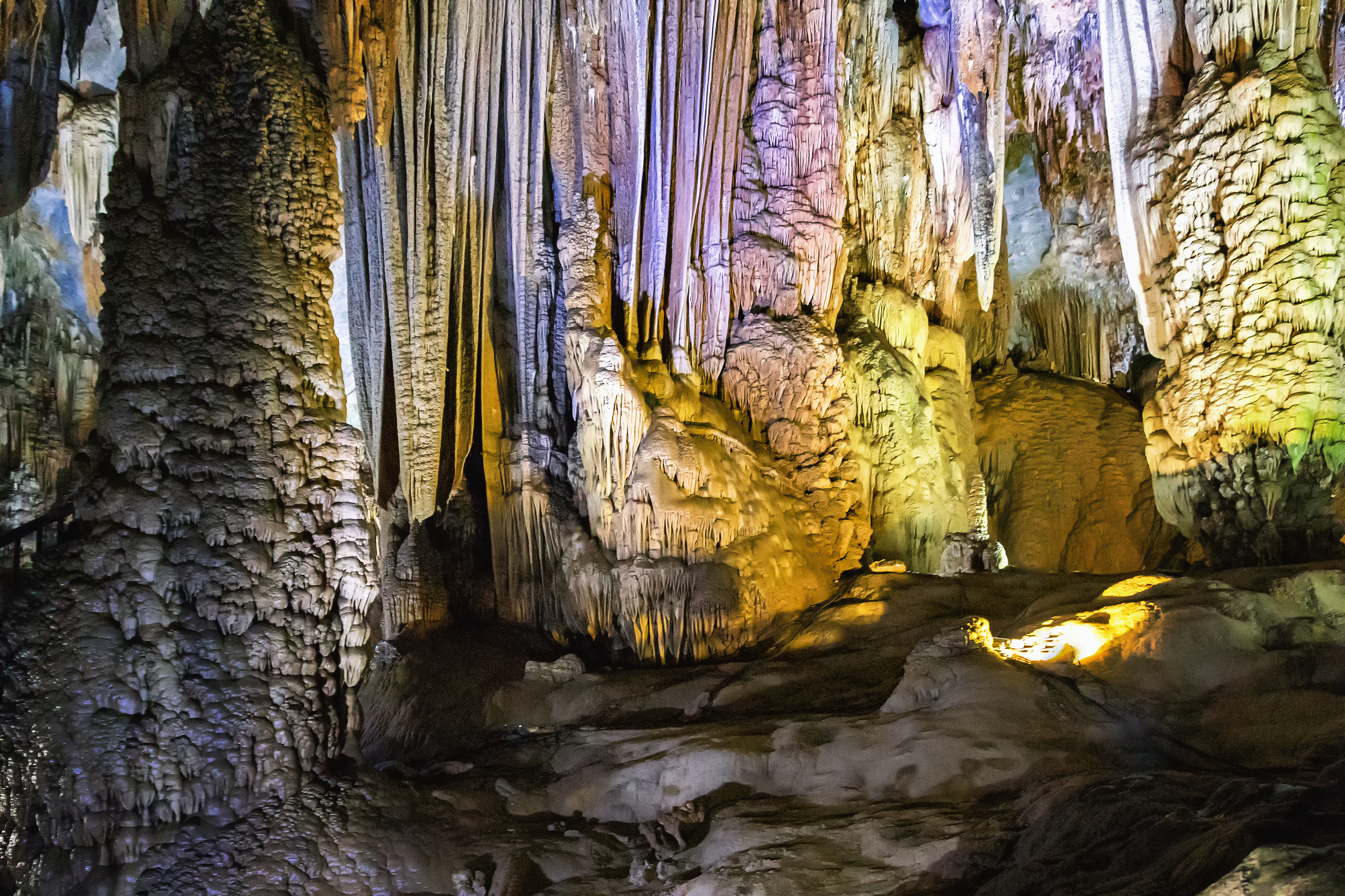 Interior de cueva con grandes formaciones de piedra caliza, estalactitas y estalagmitas, iluminado por luz artificial