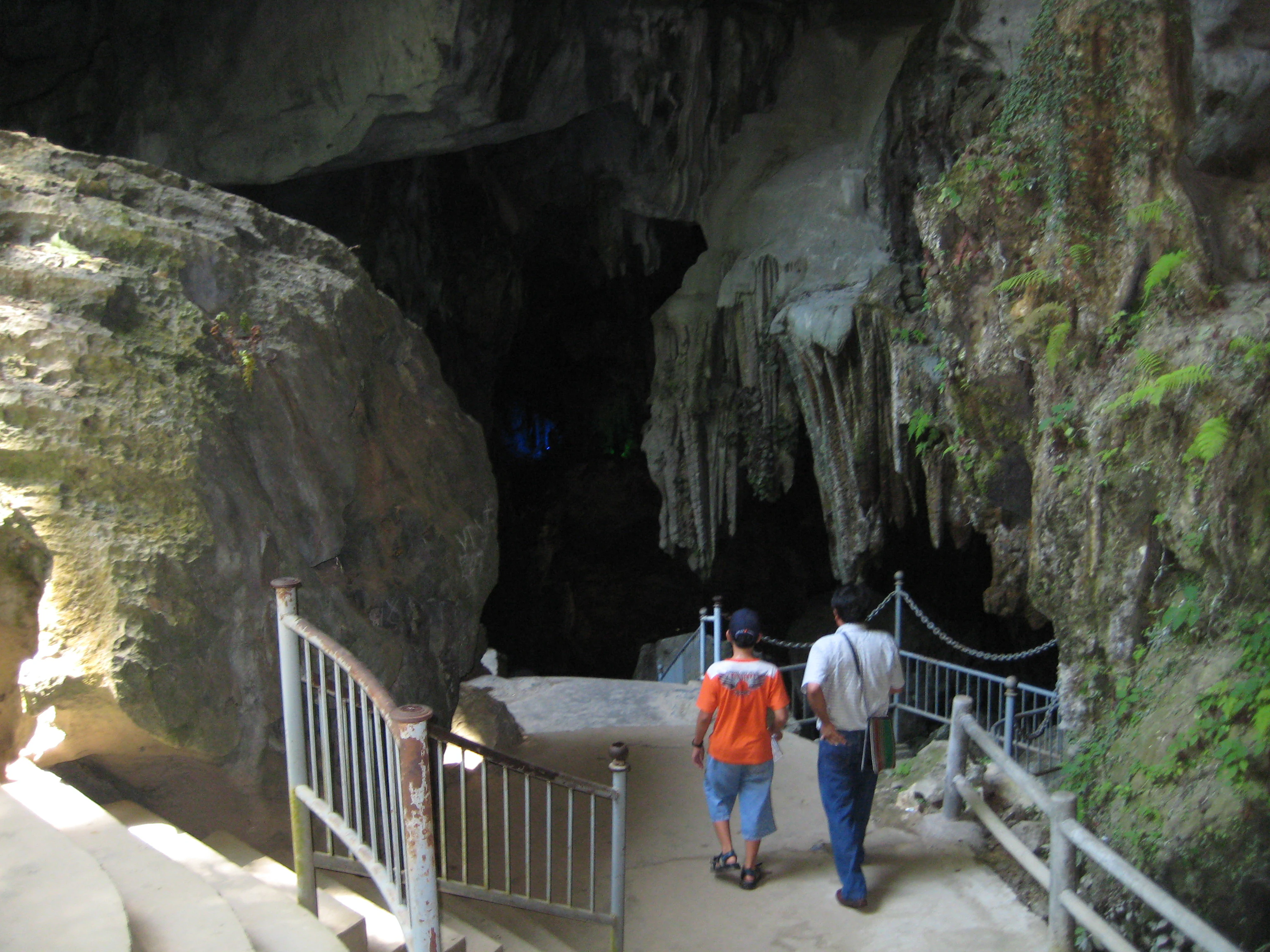 Dos personas caminan por un sendero pavimentado hacia la entrada de una cueva con paredes rocosas y barandillas metálicas.