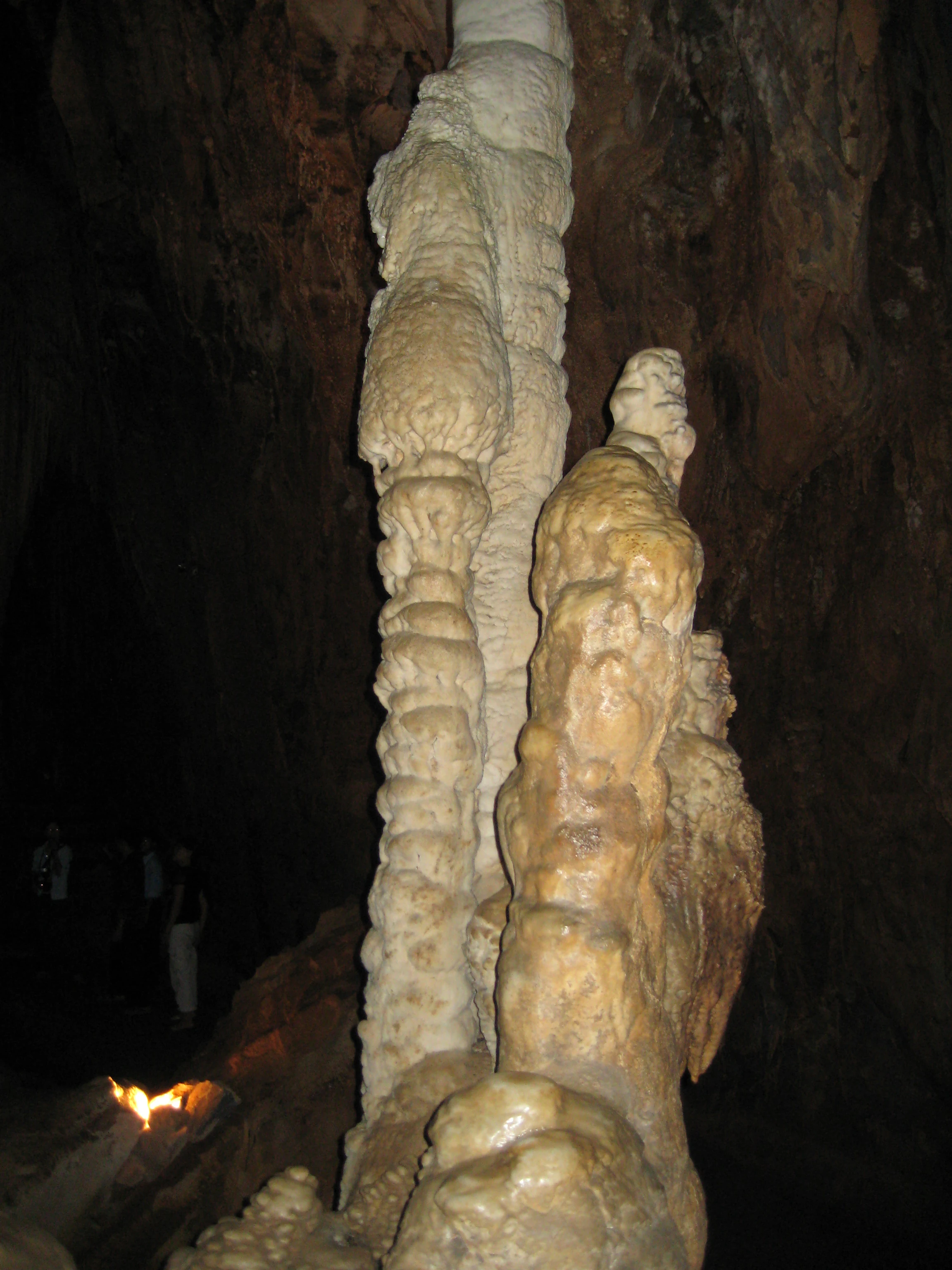 Formaciones altas de estalactitas y estalagmitas en una cueva oscura con una persona sosteniendo una fuente de luz al fondo