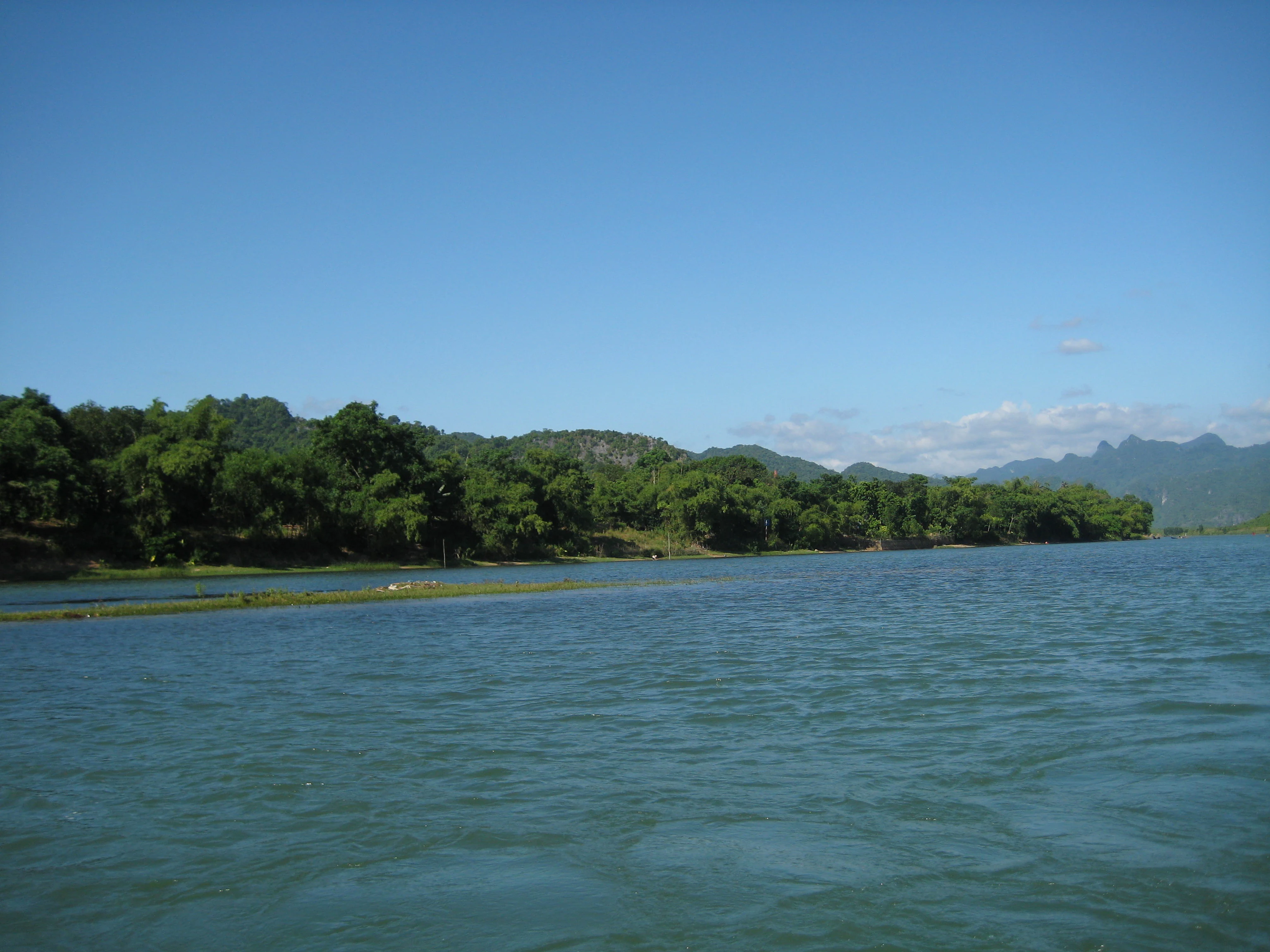Plano general de un río con orilla boscosa y colinas al fondo bajo un cielo azul despejado