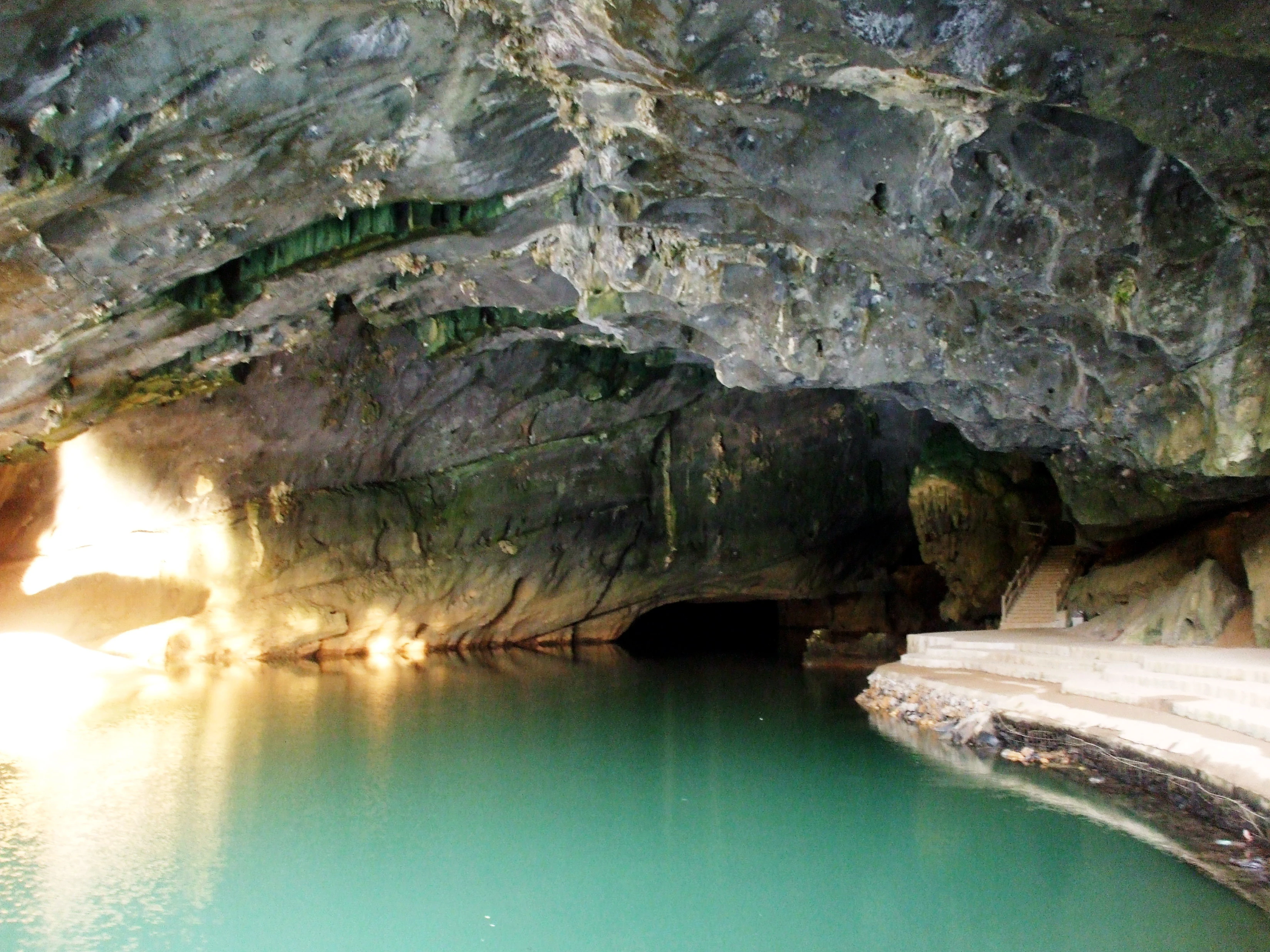 Interior de una cueva con agua de río turquesa bajo techos rocosos, iluminada por luz natural desde la entrada izquierda.