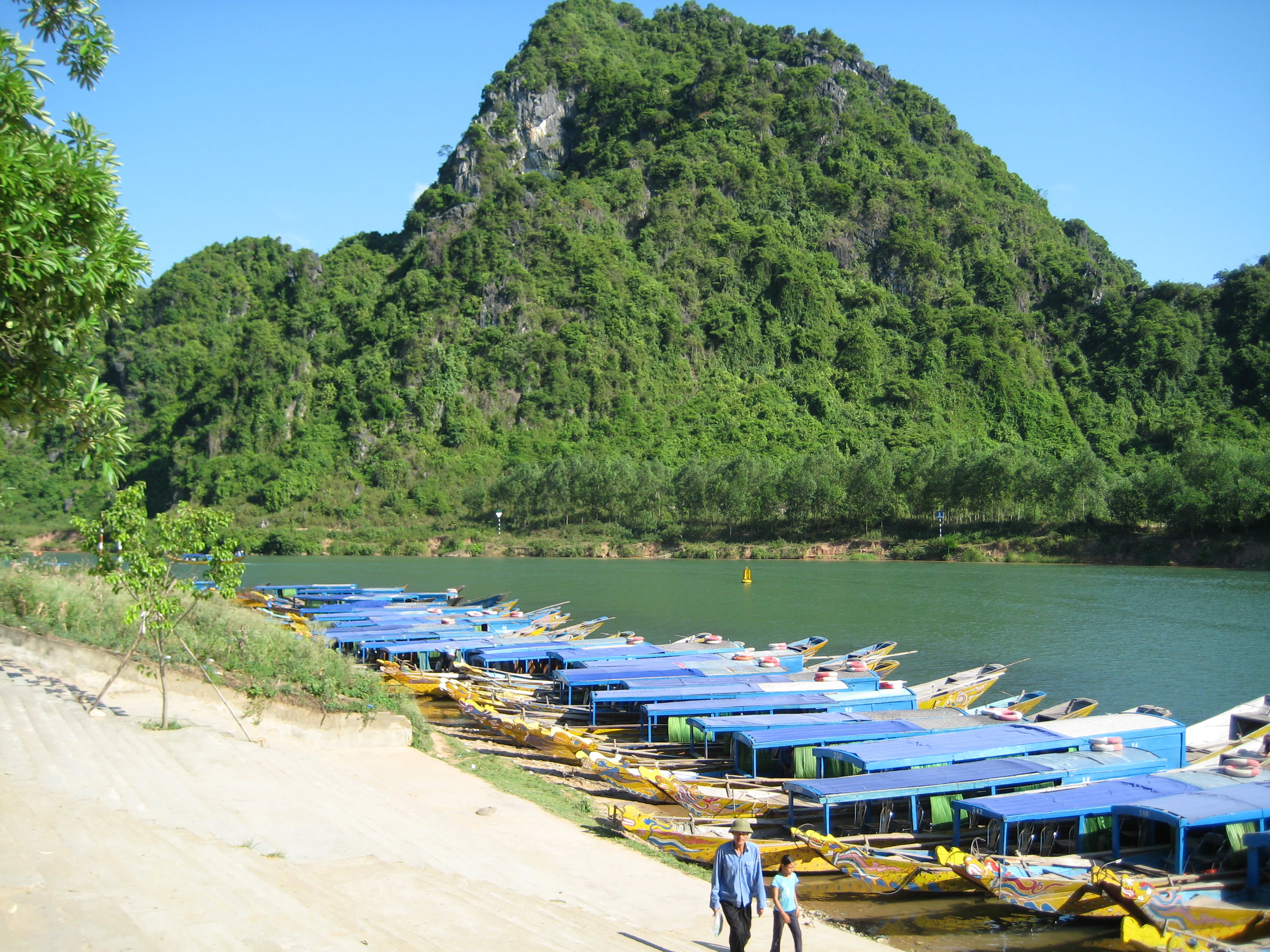 Barcas turísticas de techo azul atracadas en la orilla arenosa de un río, con una gran montaña boscosa al fondo bajo un cielo azul claro.