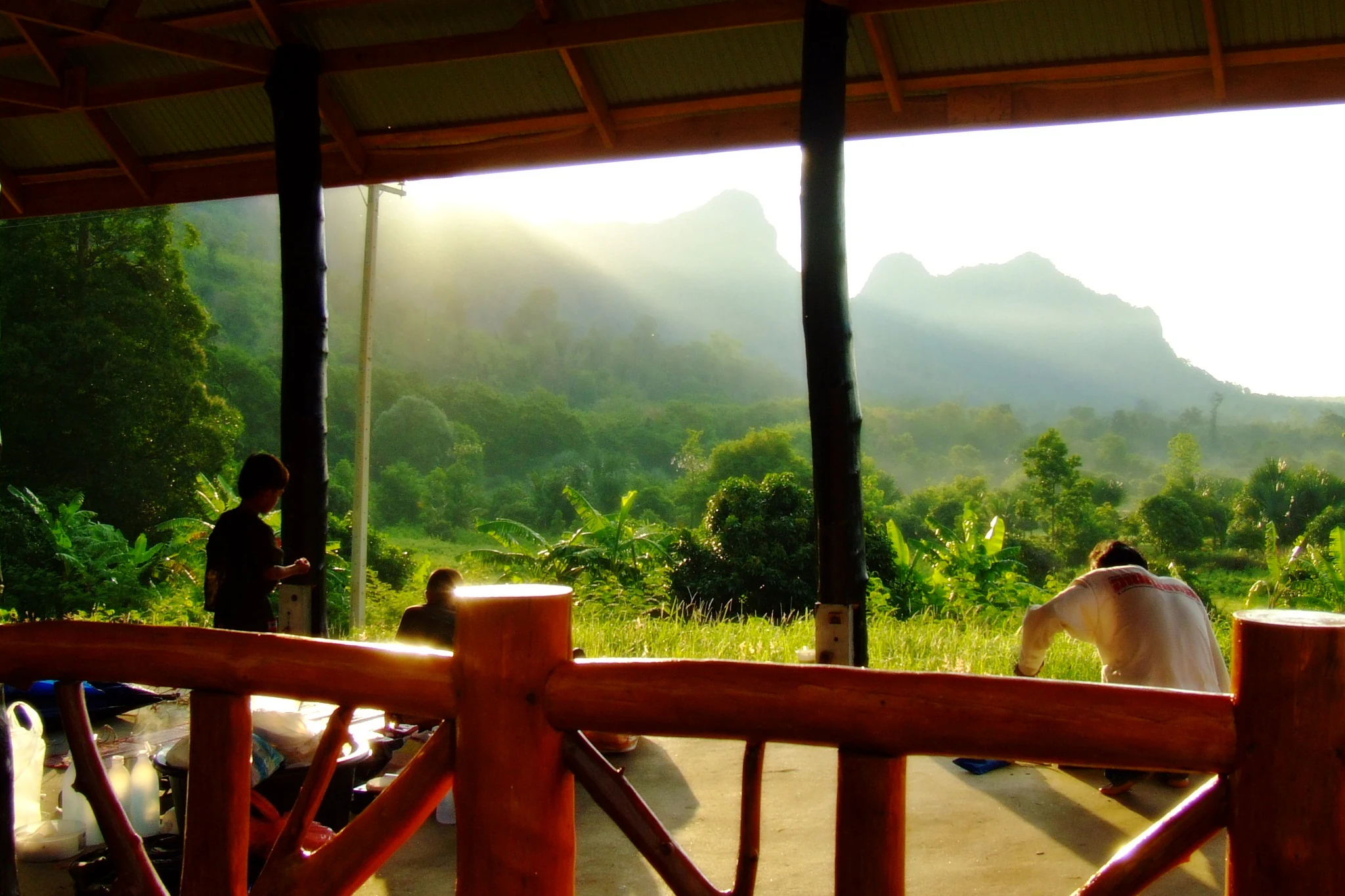 Barandilla y pilares de madera de un refugio con tres personas, con vistas a un exuberante valle verde y una cordillera con luz solar.