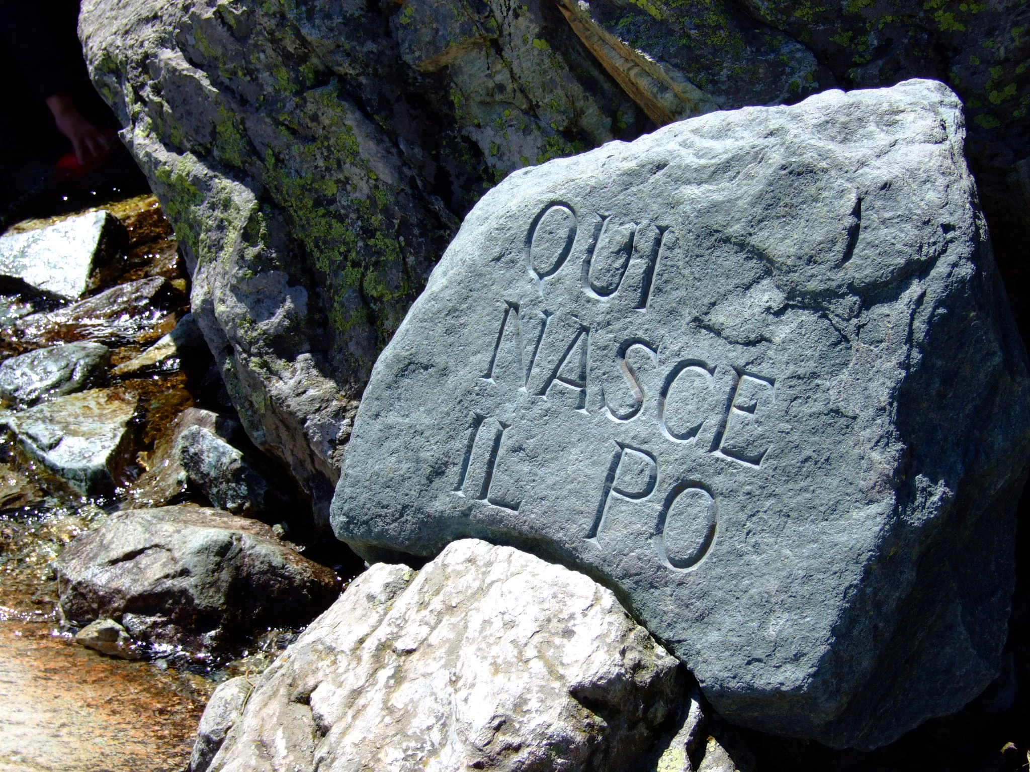 A close up view of a large grey boulder with the Italian text QUI NASCE IL PO carved into its surface, positioned next to a small mountain stream.