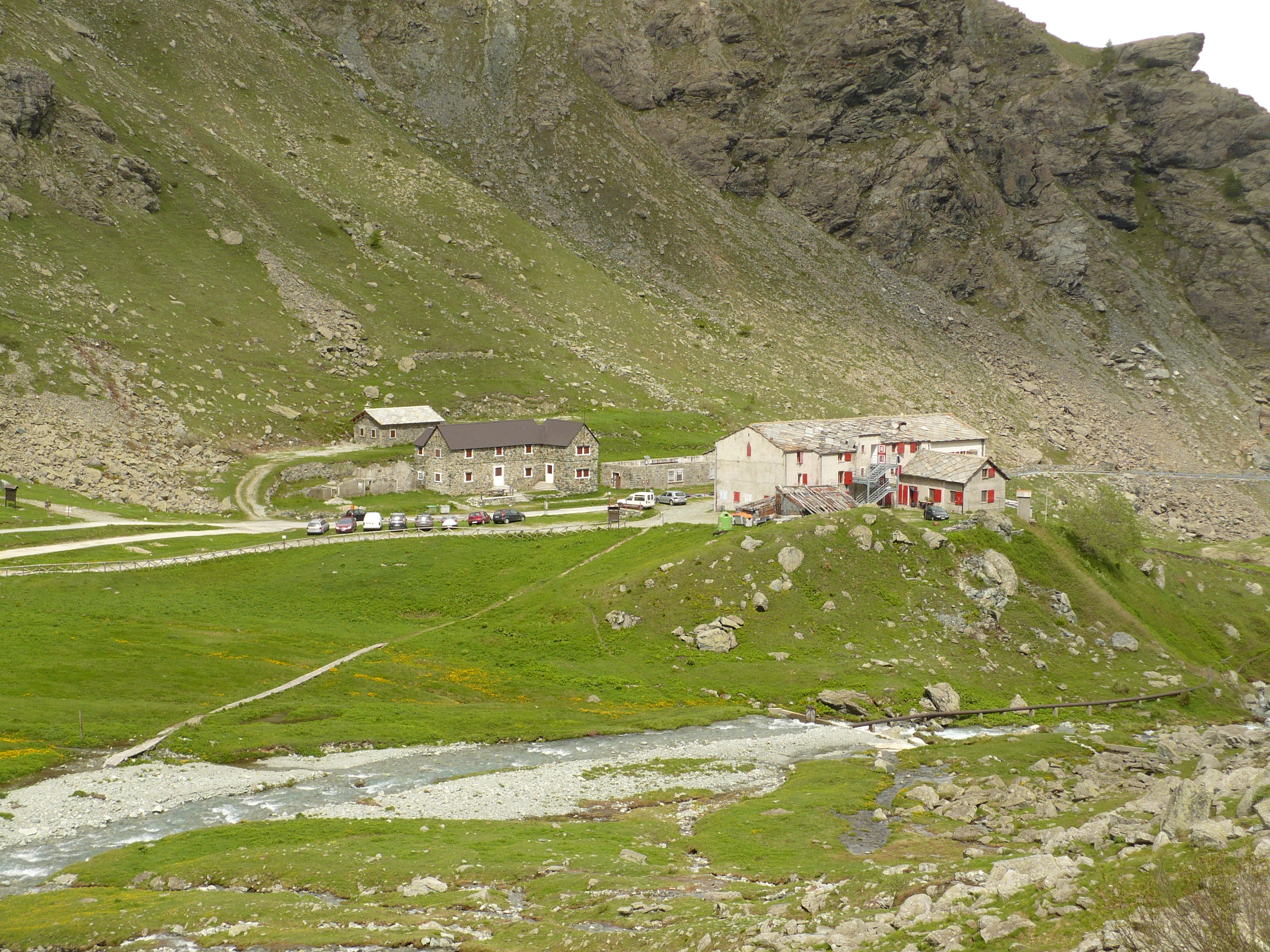 A wide view of the grassy Pian del Re plateau in a mountain valley, featuring stone buildings, a small stream, and a steep, rocky mountainside in the background.
