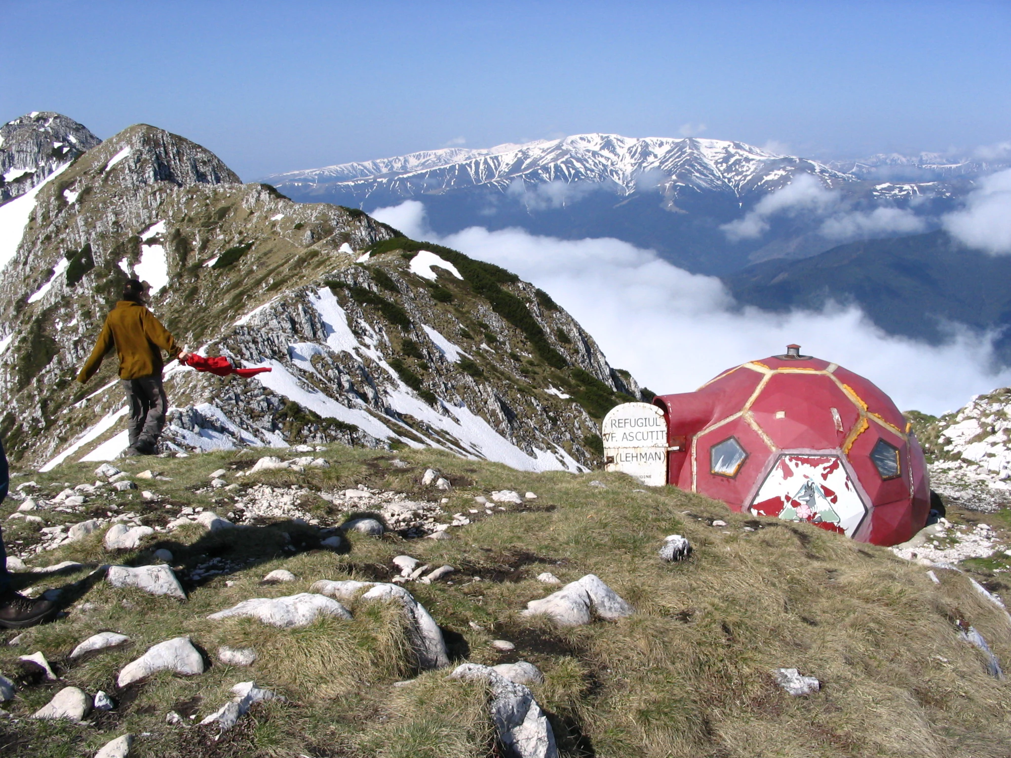 A red and yellow mountain refuge building on a rocky mountain peak with snow-capped mountains and clouds in the background, with a person in a yellow jacket standing nearby