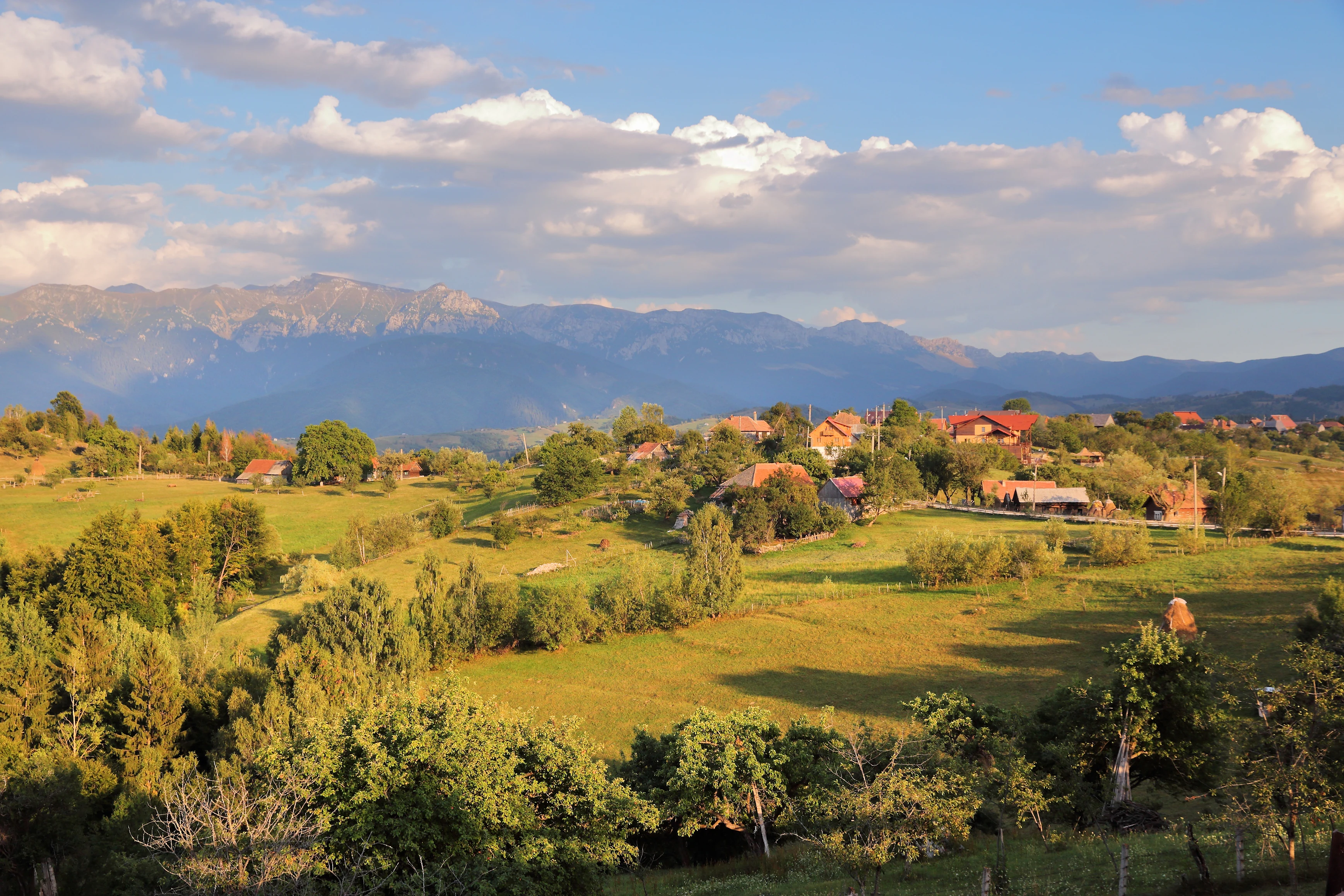 A landscape view of Magura village with Piatra Craiului mountains visible in the background, showing green fields, trees, and scattered houses under a partly cloudy sky
