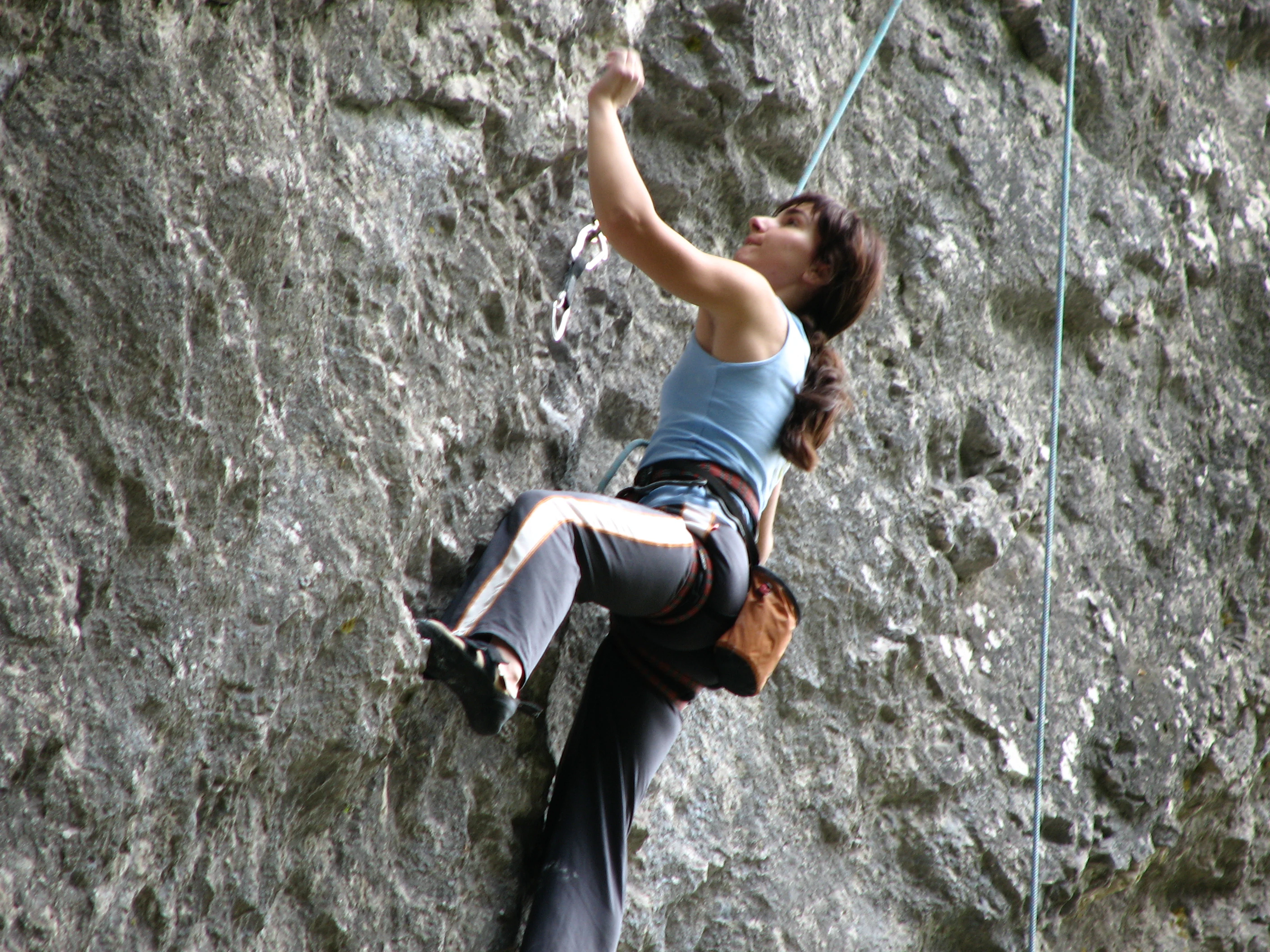 Person climbing a rock face wearing blue tank top, dark pants, harness, and climbing gear