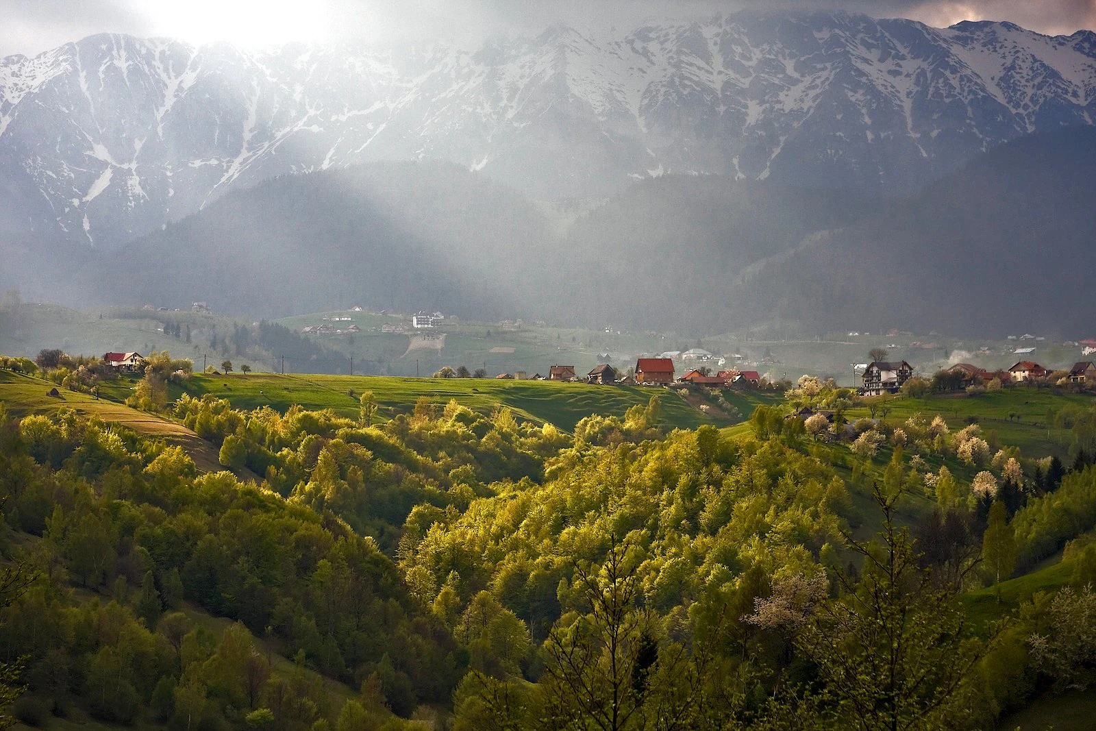 Mountain range with snow-capped peaks, rays of sunlight, green valleys, and scattered houses.