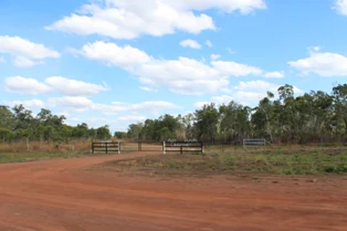 A wide dirt road leads towards a simple wooden farm-style gate, marking the entrance into a dry, grassy landscape with sparse trees under a blue sky.