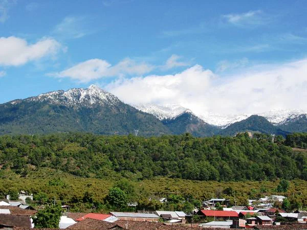 A wide shot showing a mountain range with snow-covered peaks, densely forested slopes, and a small town with red-tiled roofs in the foreground under a blue sky with light clouds.