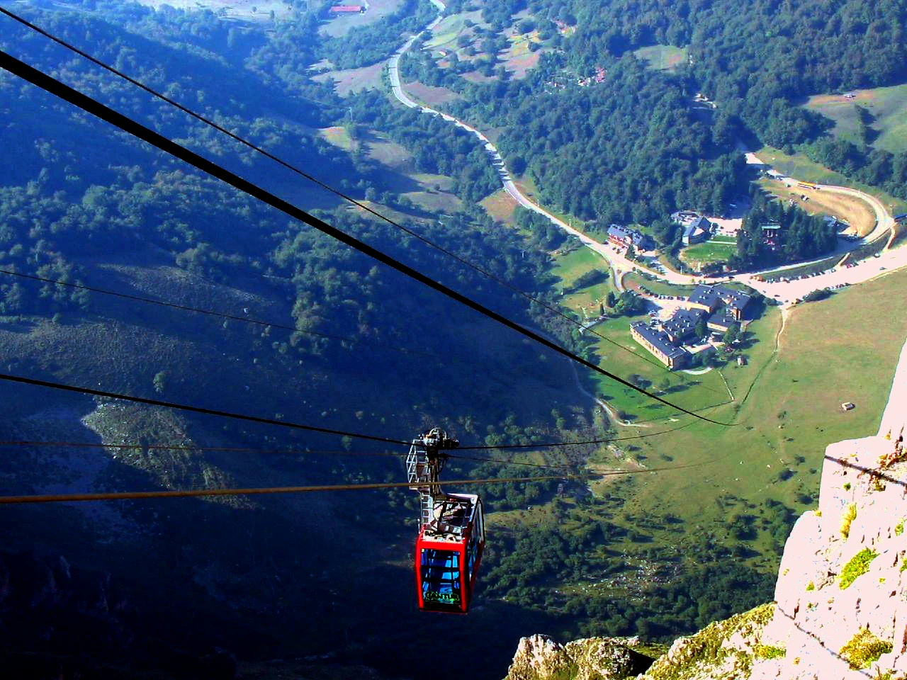 Red cable car suspended on cables above a mountainous valley with forested hills and distant buildings