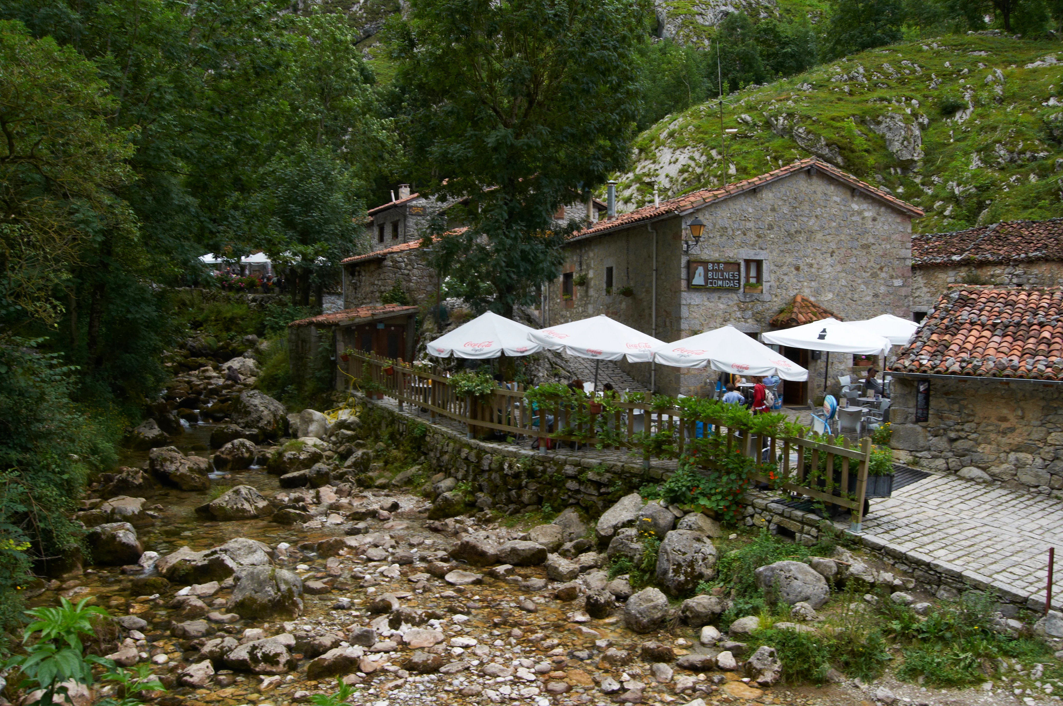 River with rocky bed, stone buildings, white umbrellas, green hills, and trees.