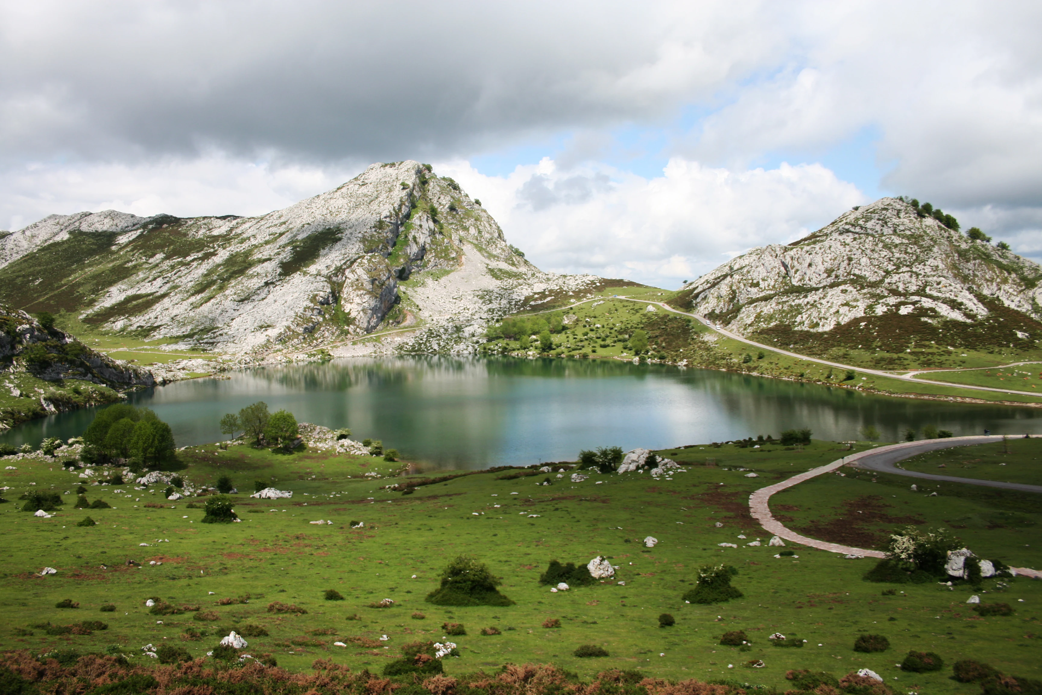 A scenic view of Lake Enol surrounded by rocky mountains and green meadows with a winding road under a partly cloudy sky