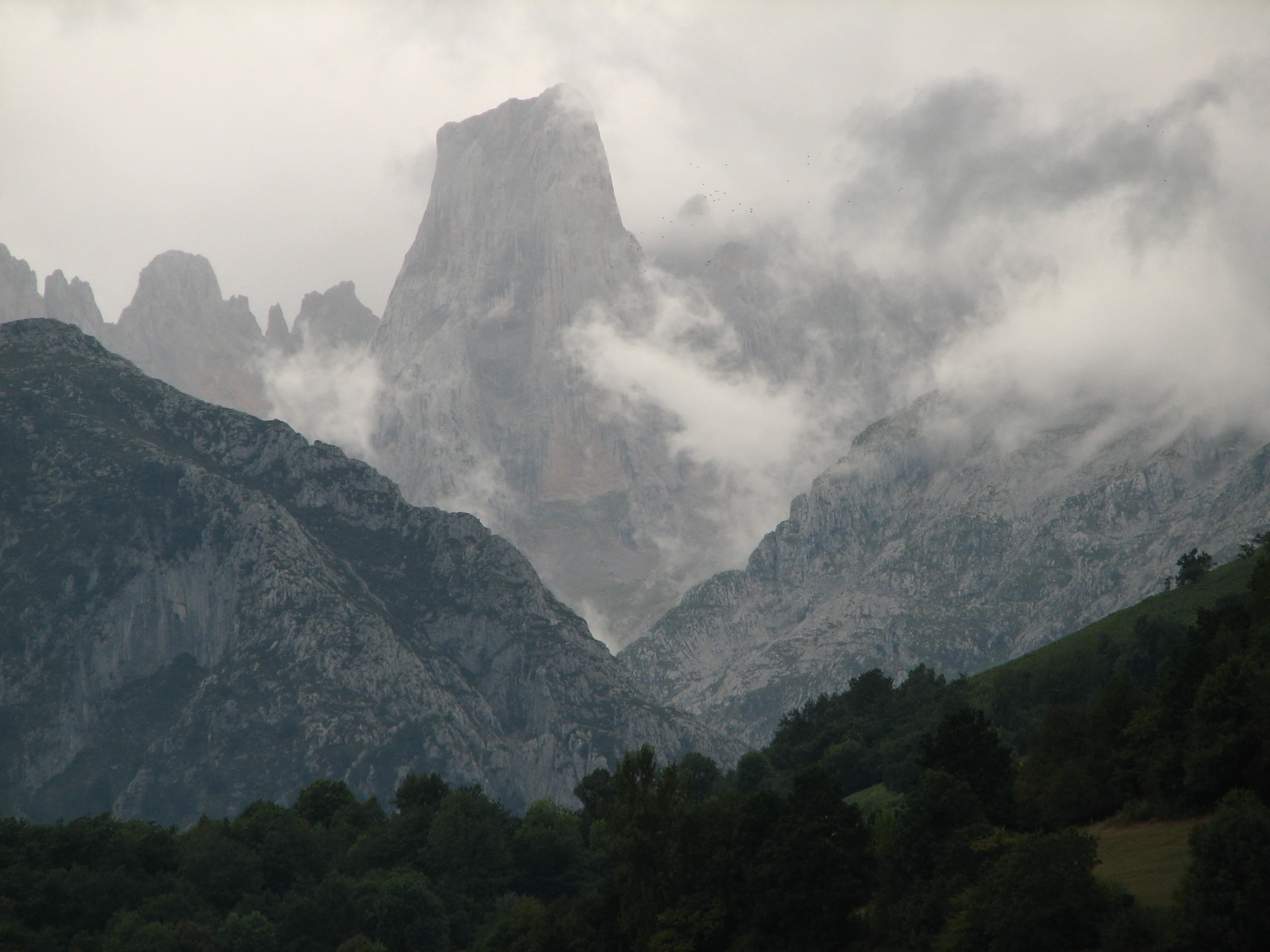 Mountainous landscape featuring a prominent rocky peak partially obscured by clouds, with forested areas in the foreground and rugged rock formations in the background