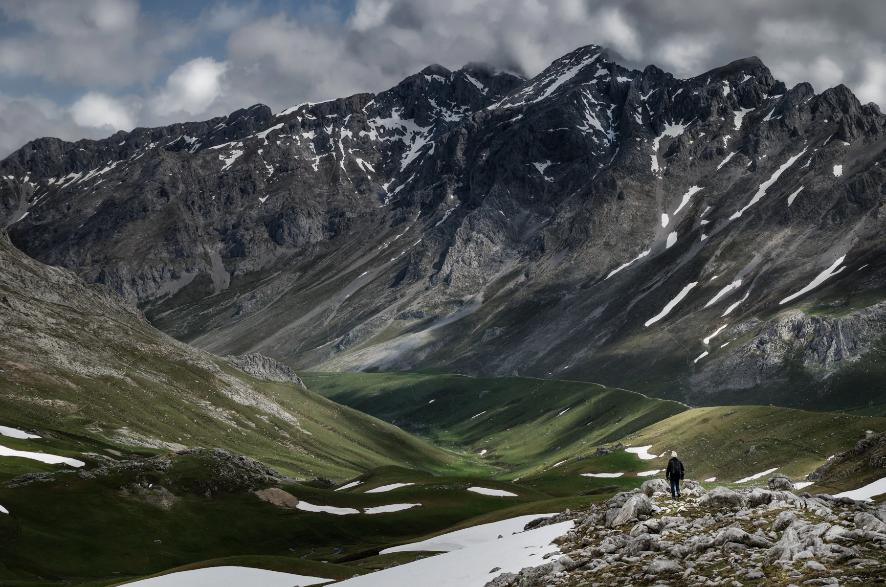 A person standing on rocky terrain with patches of snow, green valleys, and mountain peaks in the background