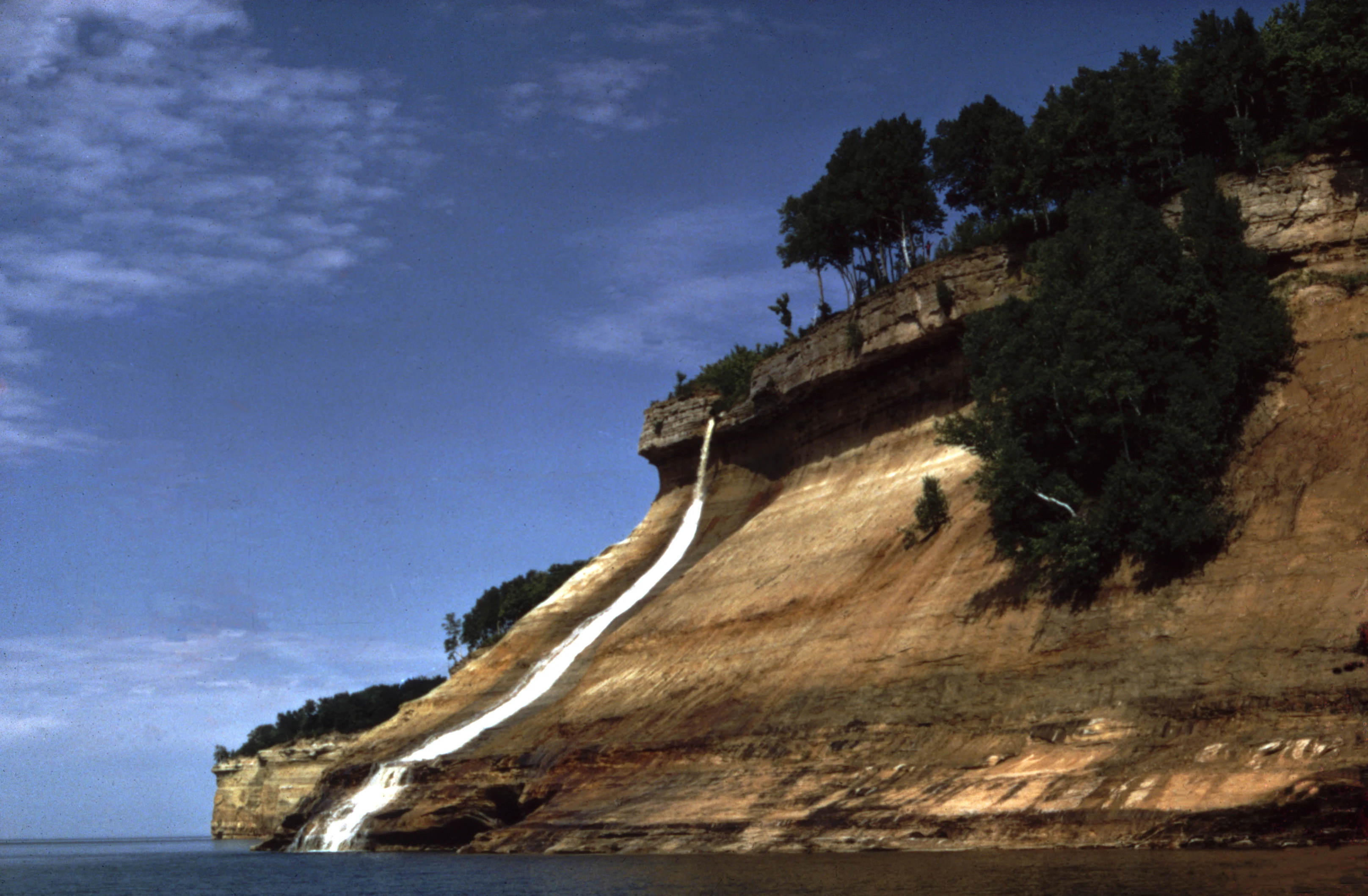 Una cascada se precipita por el borde de un alto acantilado de arenisca en capas hacia el lago Superior, rodeada de árboles en la parte superior.