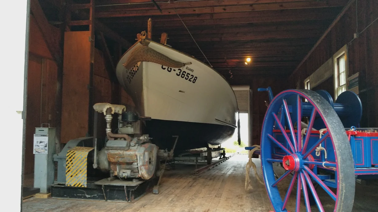 Interior de una casa de botes histórica de madera que contiene un bote salvavidas blanco de la Guardia Costera etiquetado CG-36528 y un carro de rescate azul con ruedas grandes.