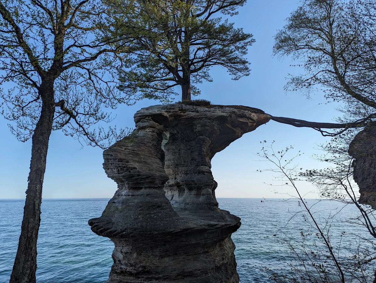 Una gran formación rocosa de arenisca, Chapel Rock, se alza contra el telón de fondo del lago Superior, con un árbol creciendo en su cima y un puente de raíces expuestas que se extiende hacia el acantilado adyacente.