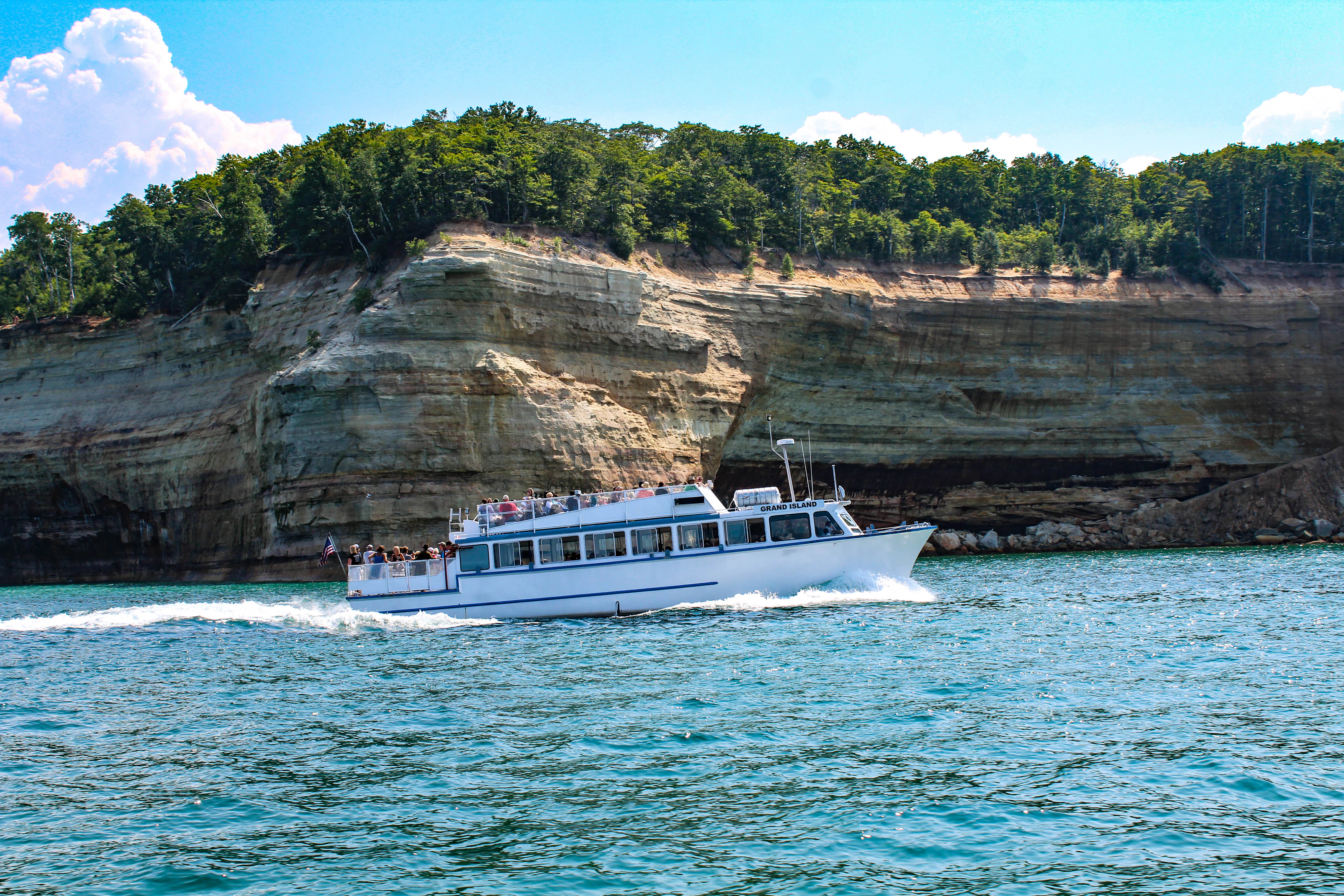 Un barco turístico blanco con pasajeros visibles en la cubierta navega por aguas azules frente a un acantilado alto y estratificado de arenisca coronado por un bosque verde.