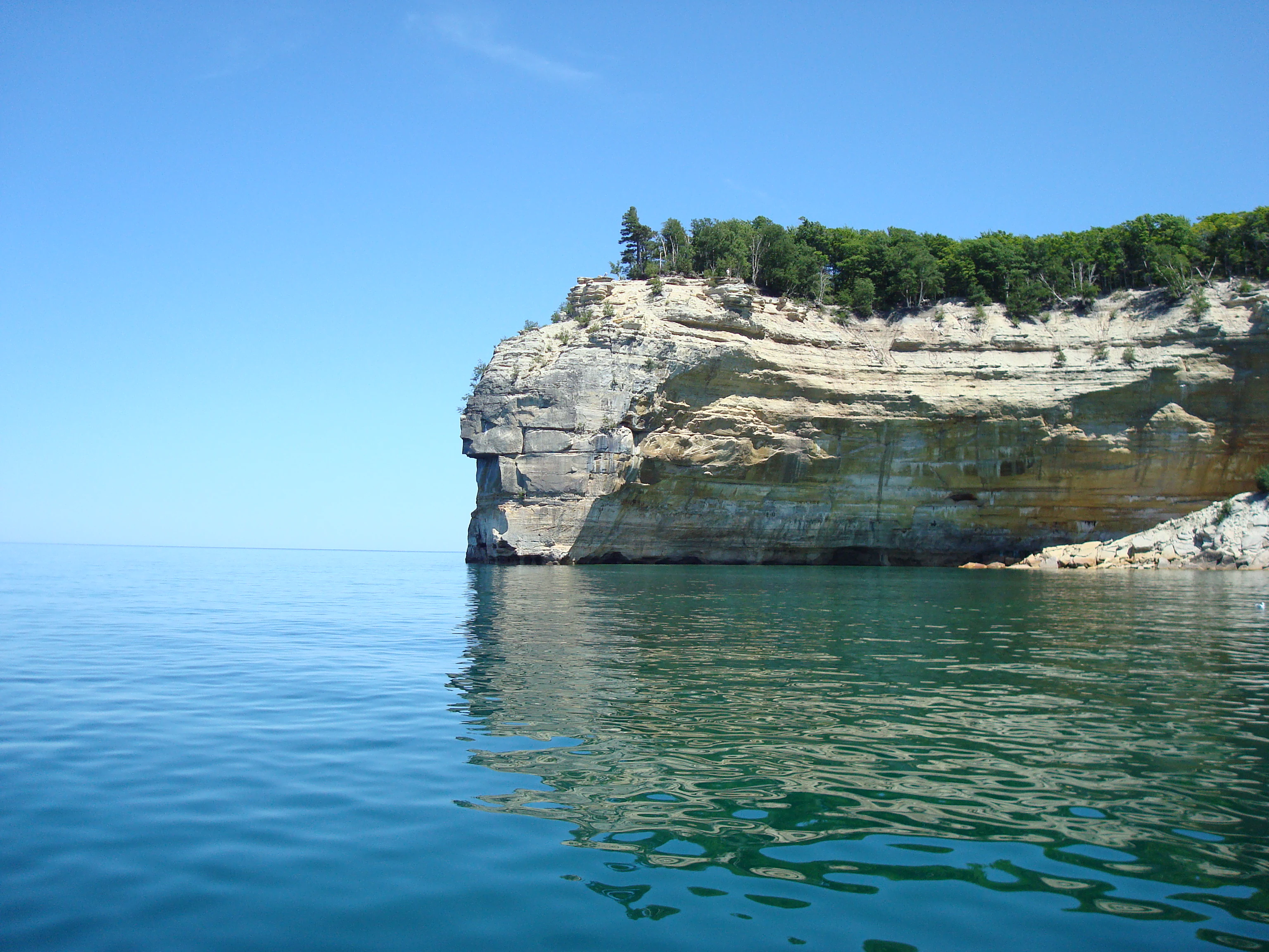 Una formación de acantilados de arenisca que se asemeja a un perfil humano, conocida como Indian Head, se adentra en las tranquilas aguas azules del lago Superior, con un dosel forestal en la cima de la roca.