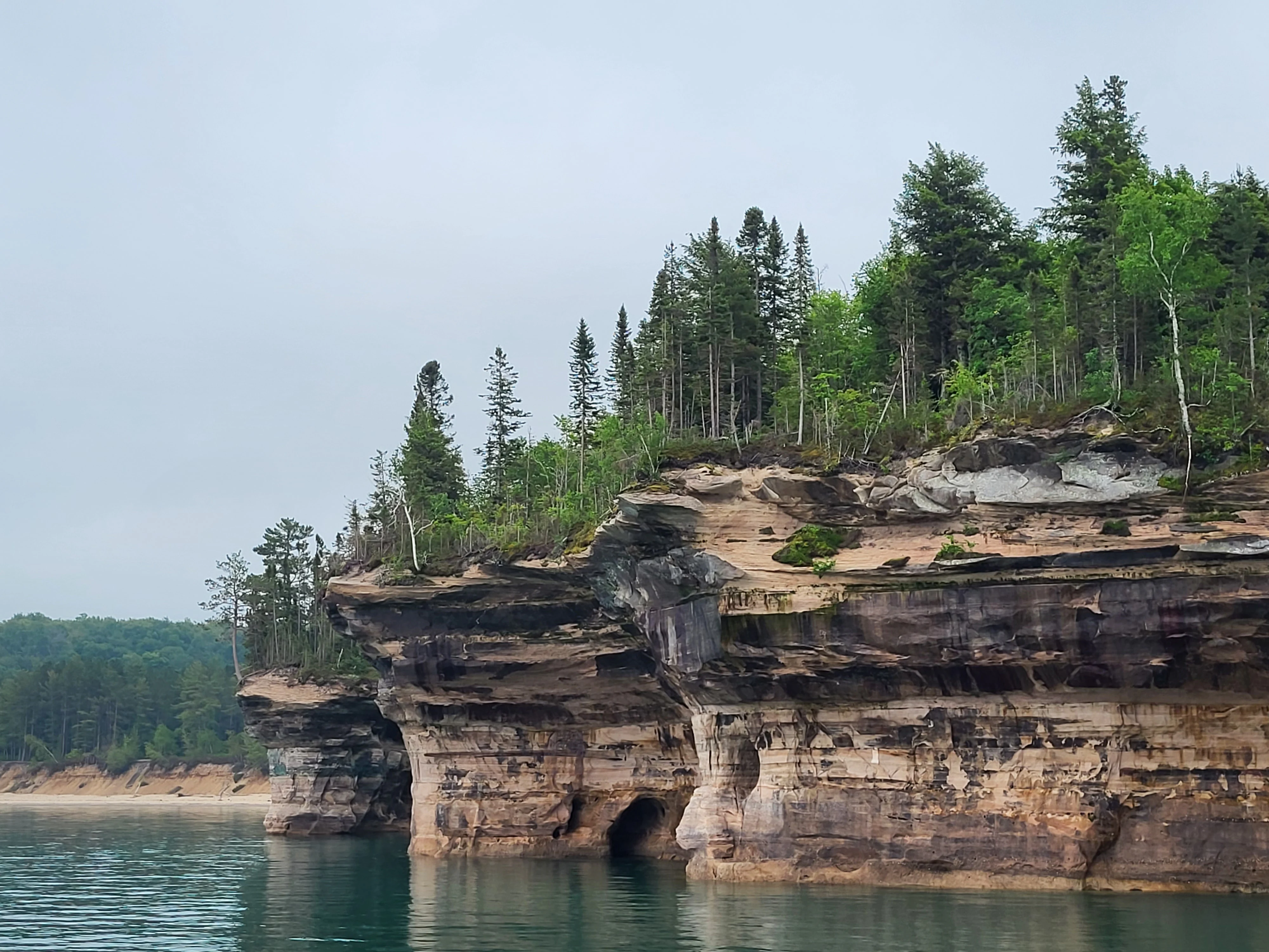 Una vista amplia de acantilados de arenisca estratificados y coloridos que se elevan directamente desde el lago Superior, coronados por un denso bosque de árboles de hoja perenne y caducifolia bajo un cielo nublado.