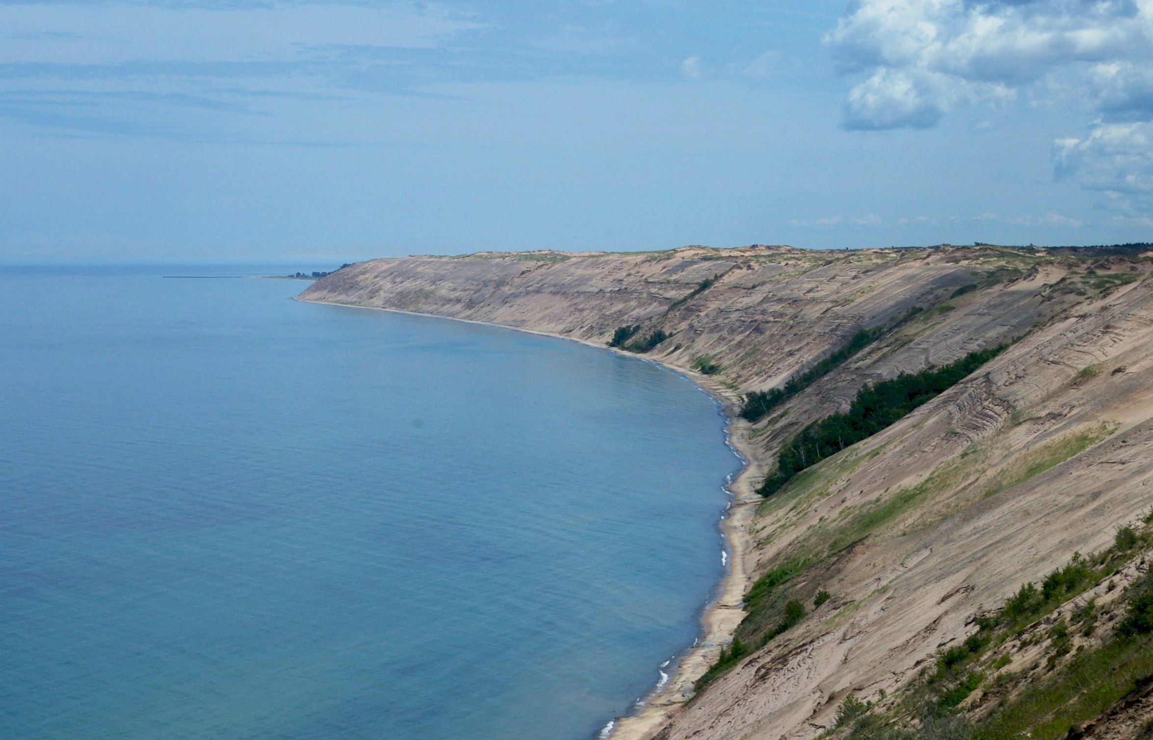 Una vista de gran angular a lo largo de la costa, donde enormes dunas de arena y acantilados se encuentran con las aguas azules del lago Superior bajo un cielo parcialmente nublado.