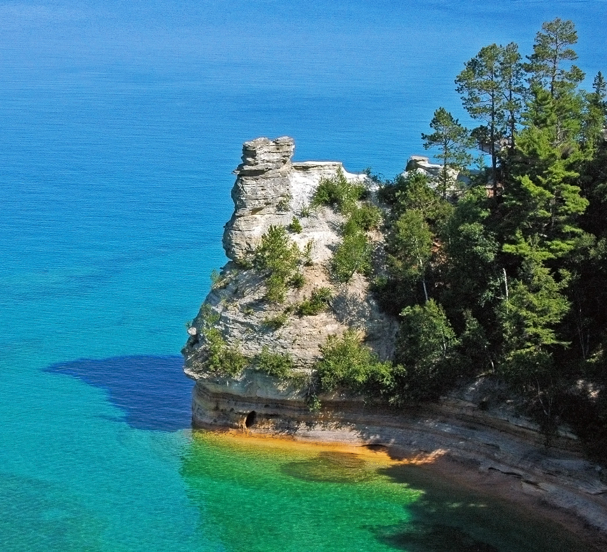 Una vista en ángulo alto de la formación rocosa de arenisca Miners Castle que se extiende hacia las brillantes aguas turquesas del lago Superior, rodeada de pinos verdes.