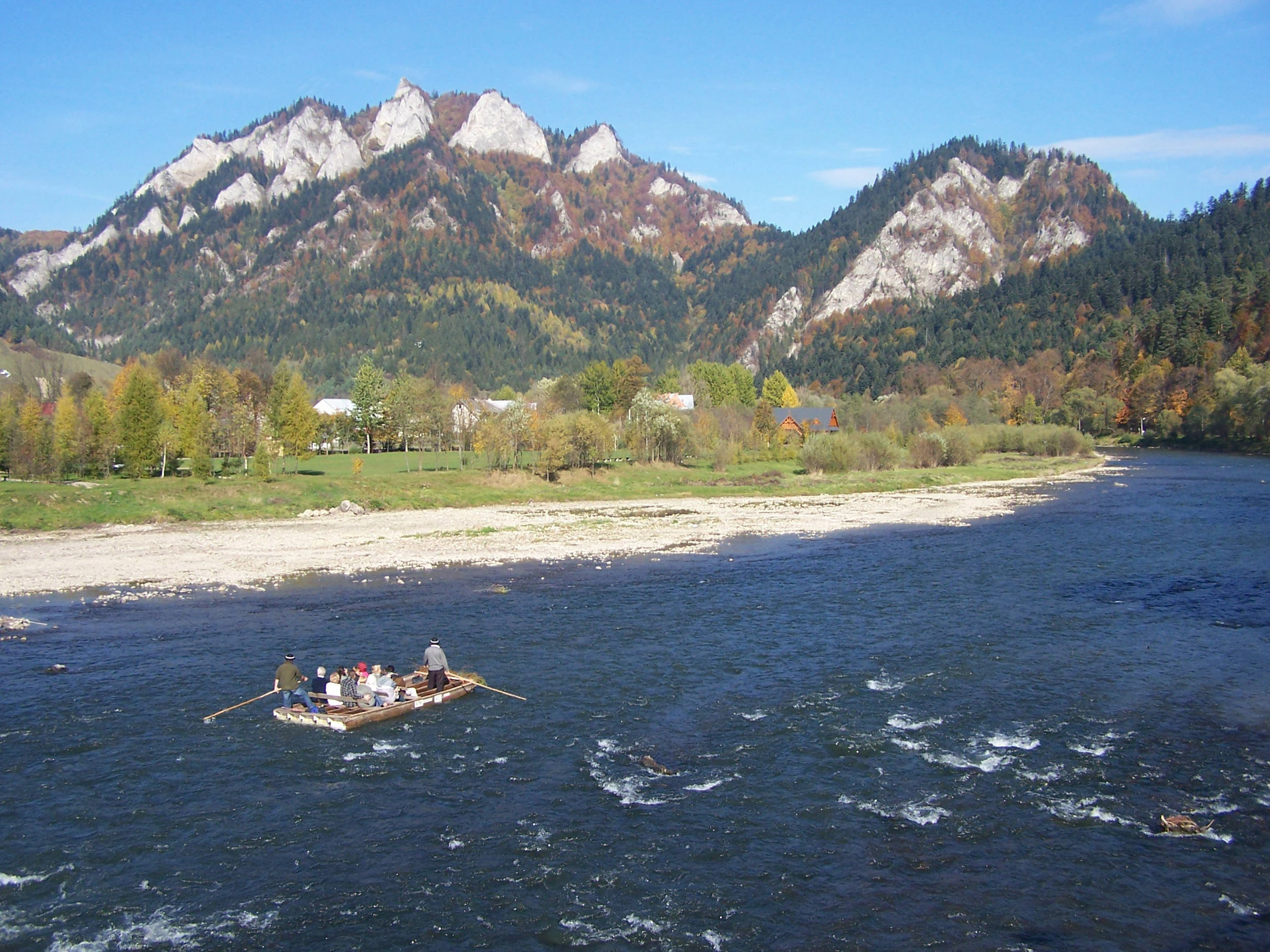 Un grupo de personas en una balsa de madera navega por el río Dunajec, con picos de piedra caliza boscosos al fondo durante el otoño.