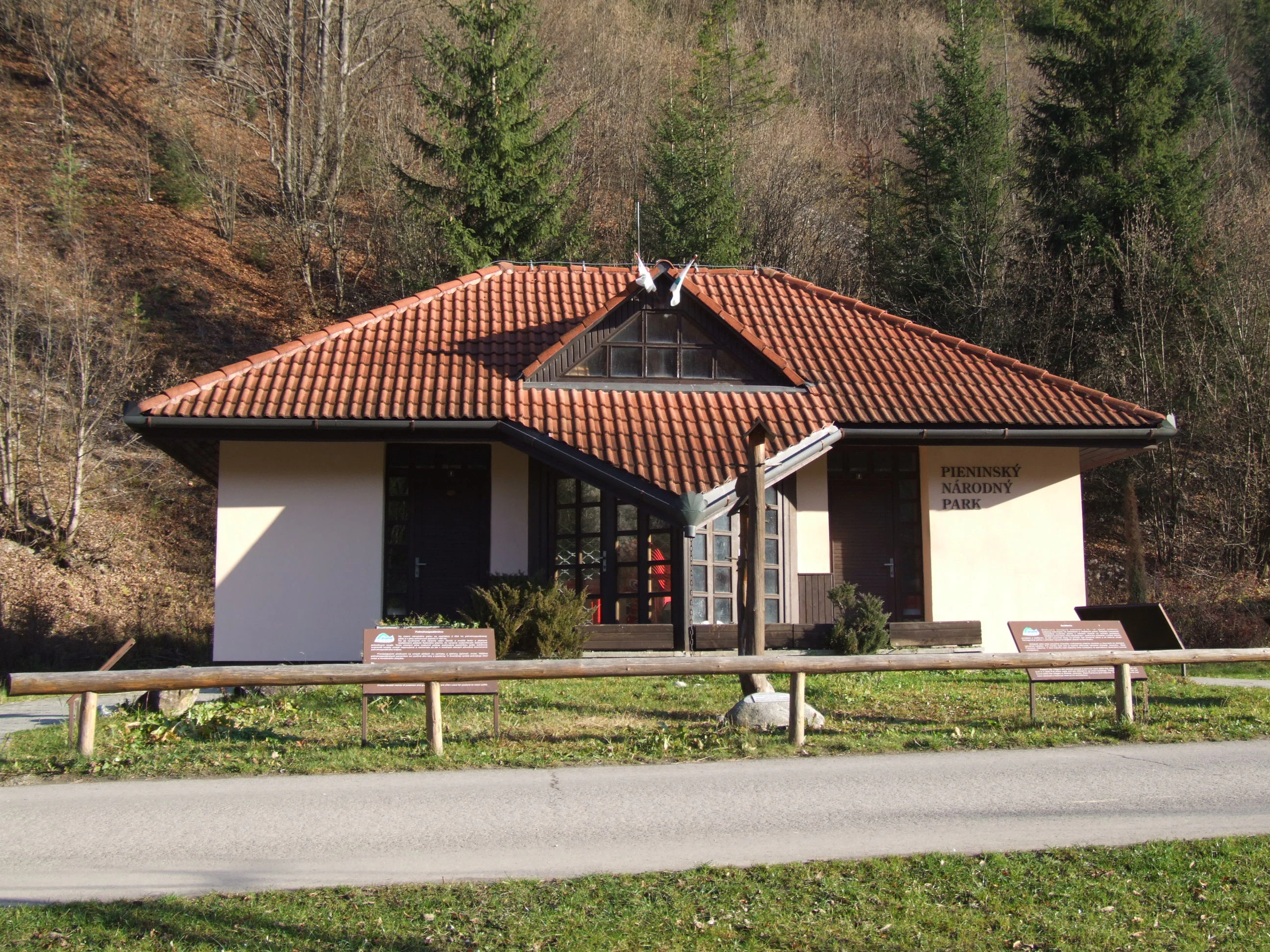 A white building with a red-tiled roof and wooden fence, situated in a forested area with trees and grass