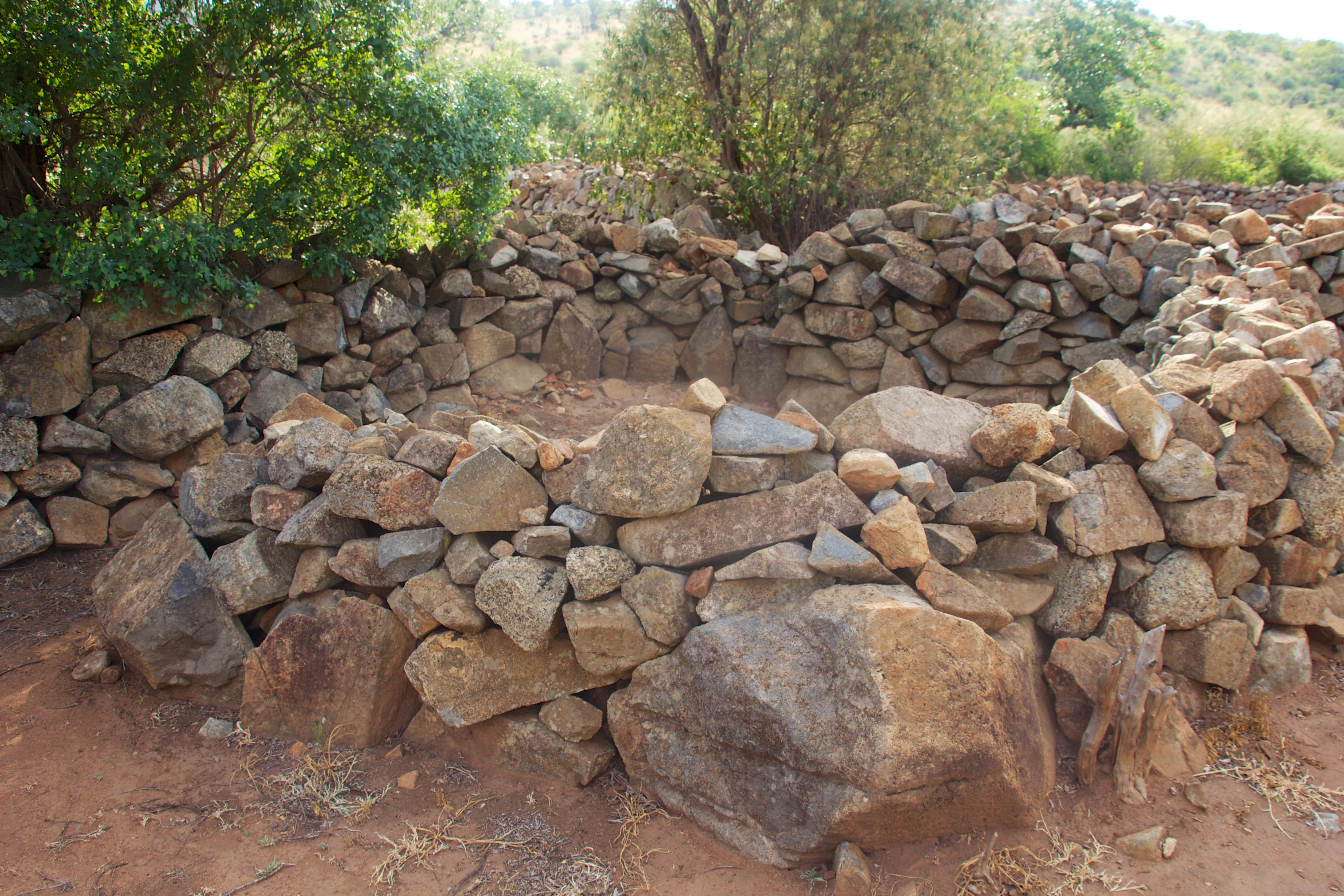 Ancient circular stone wall remains constructed from stacked rocks nestled among sparse trees and bushes.