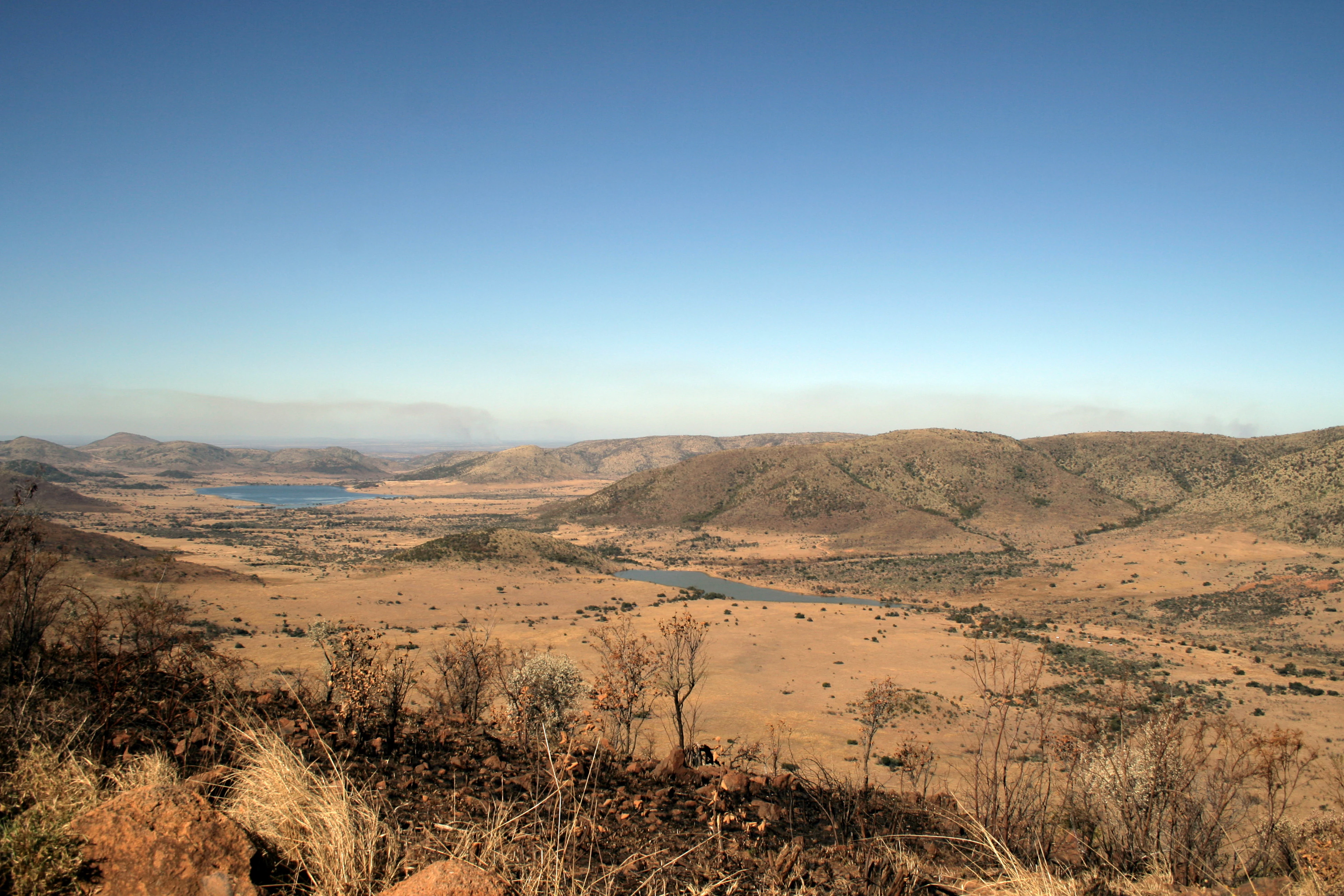 A wide, high-angle landscape shot showing arid, grassy savanna plains and distant water bodies surrounded by low, rolling hills under a clear blue sky.