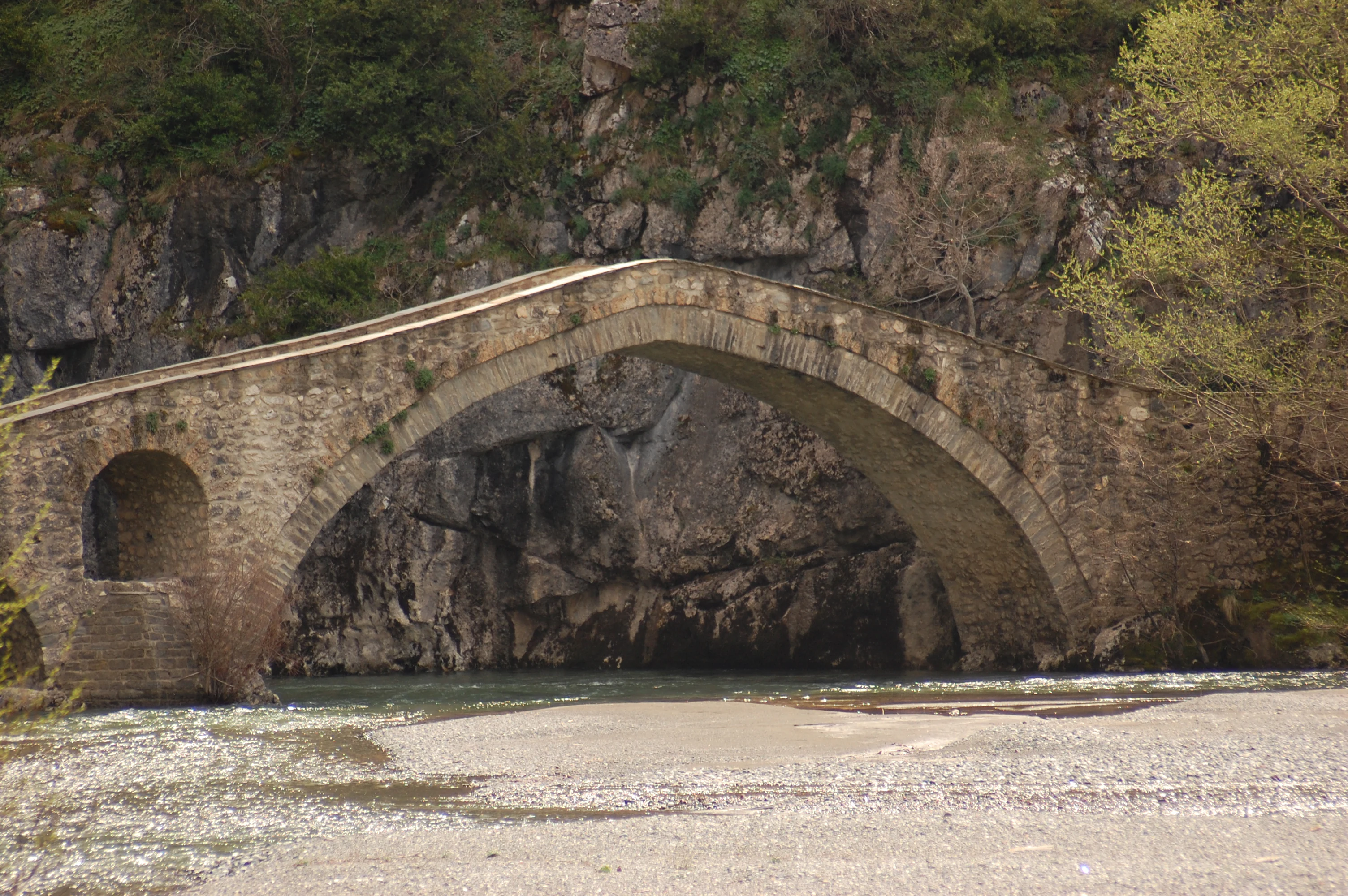 Stone arch bridge over a river with rocky cliffs and vegetation in the background