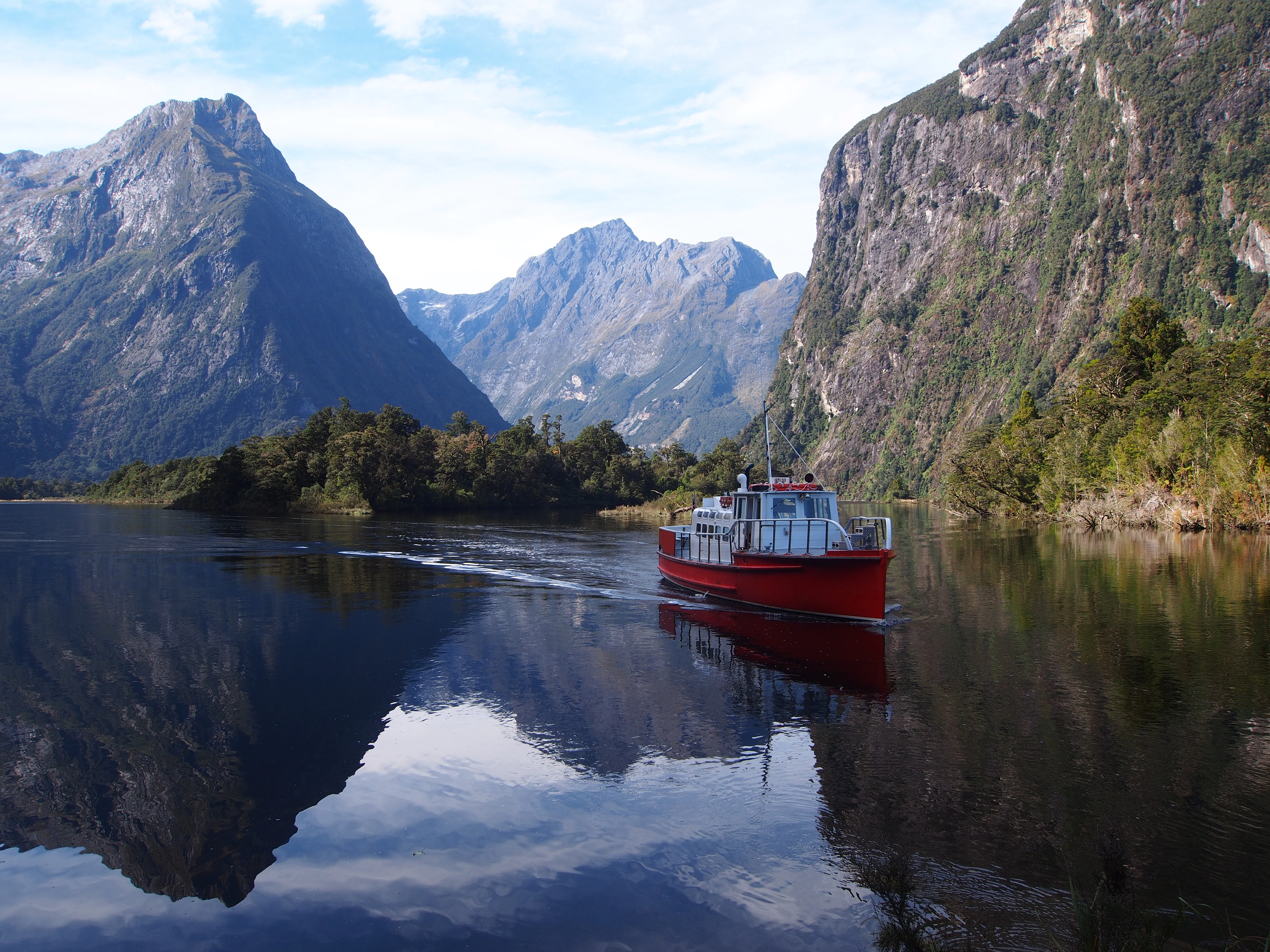 A bright red boat cruises on the calm, reflective waters of a fjord with steep, forested mountains on both sides.