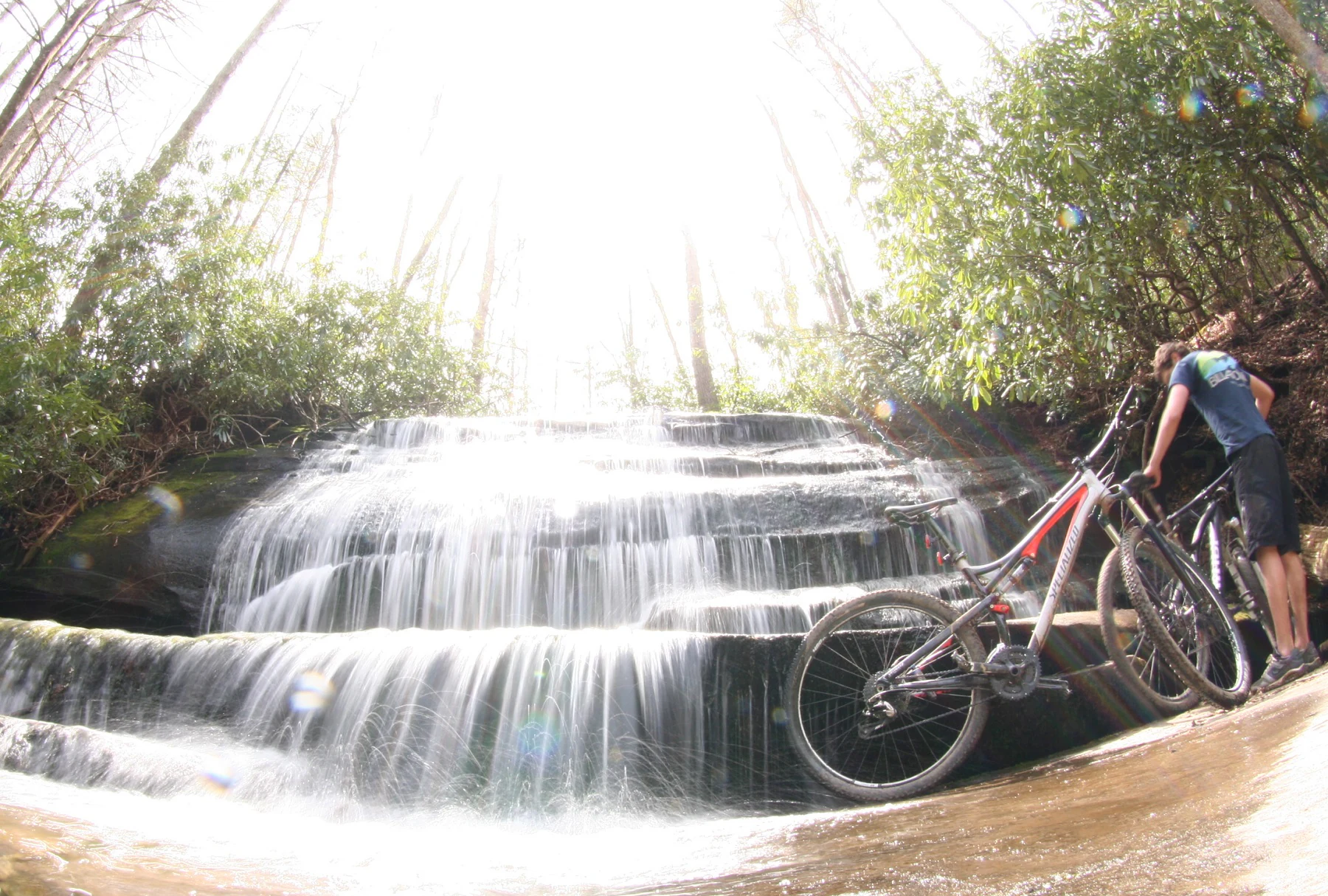 A mountain biker stands beside their bicycle on a trail next to a small, tiered waterfall in a forest setting.