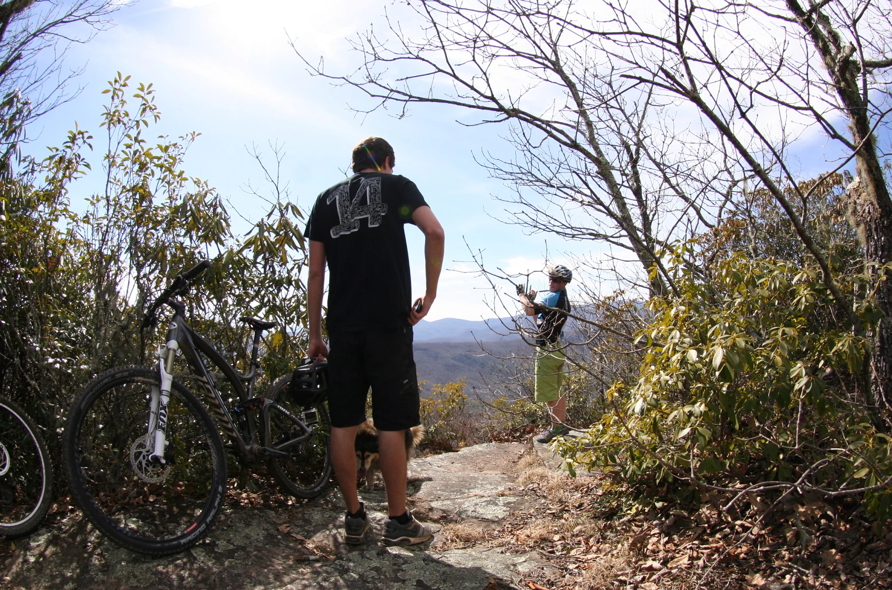 Two mountain bikers standing on a rocky trail overlook, with one standing in the foreground and another looking at a view of distant mountain ridges.