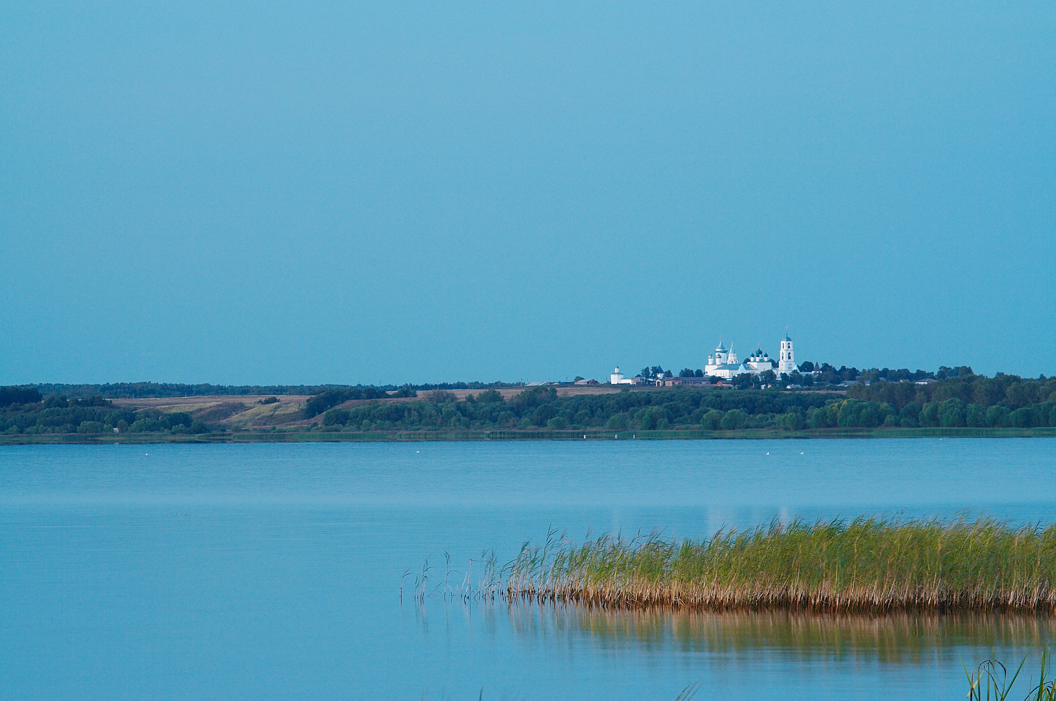 Calm blue lake with tall reeds in foreground and white monastery on distant hill under clear blue sky