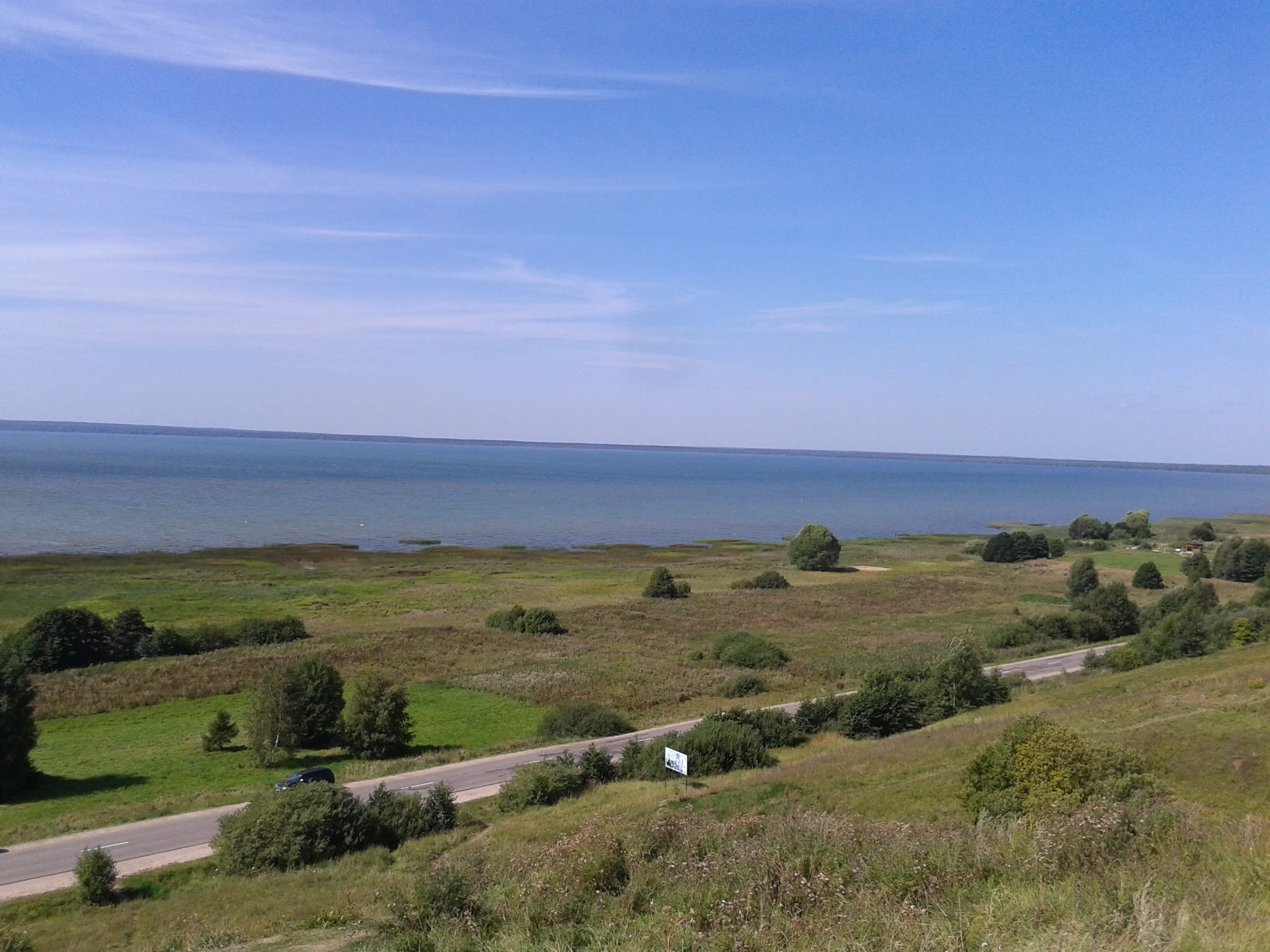 A wide-angle landscape view looking out over a large lake with grassy plains and a road in the foreground under a clear blue sky.