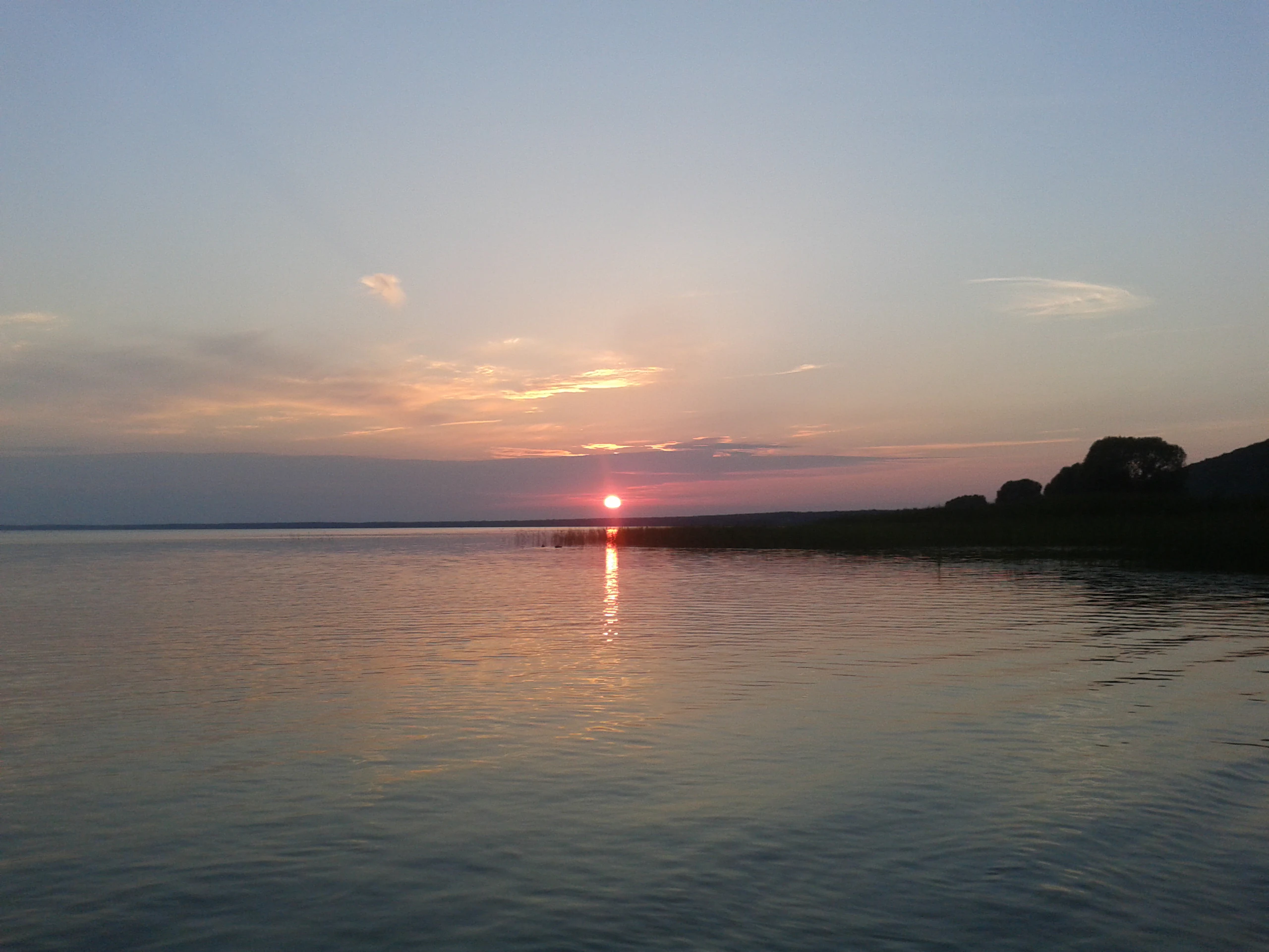A wide shot of a calm lake at sunset, with the sun low on the horizon, casting a soft reflection on the water and silhouetting a low grassy shoreline.