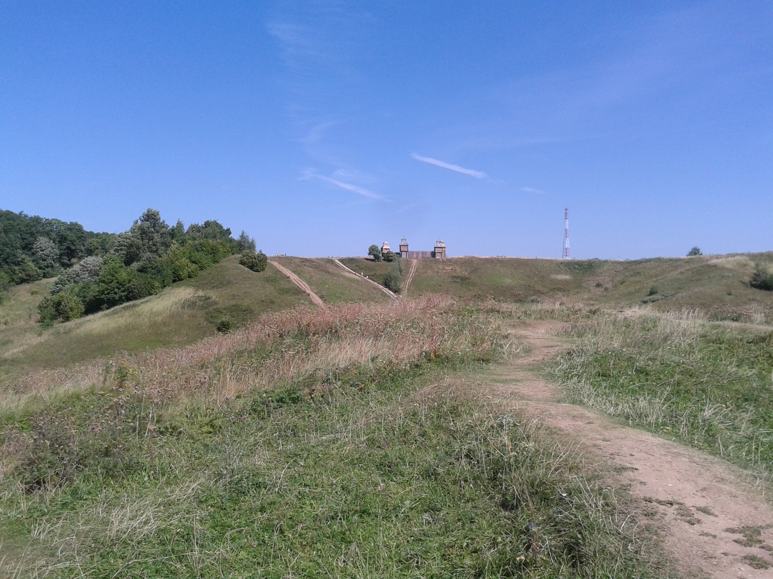A dirt hiking trail leads across a grassy hill under a clear blue sky, with a small building and a tall communication tower visible in the distance on a ridgeline.