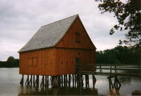 A brown timber stilt house stands on wooden pilings above the water of a pond, connected to the shore by a short wooden footbridge.