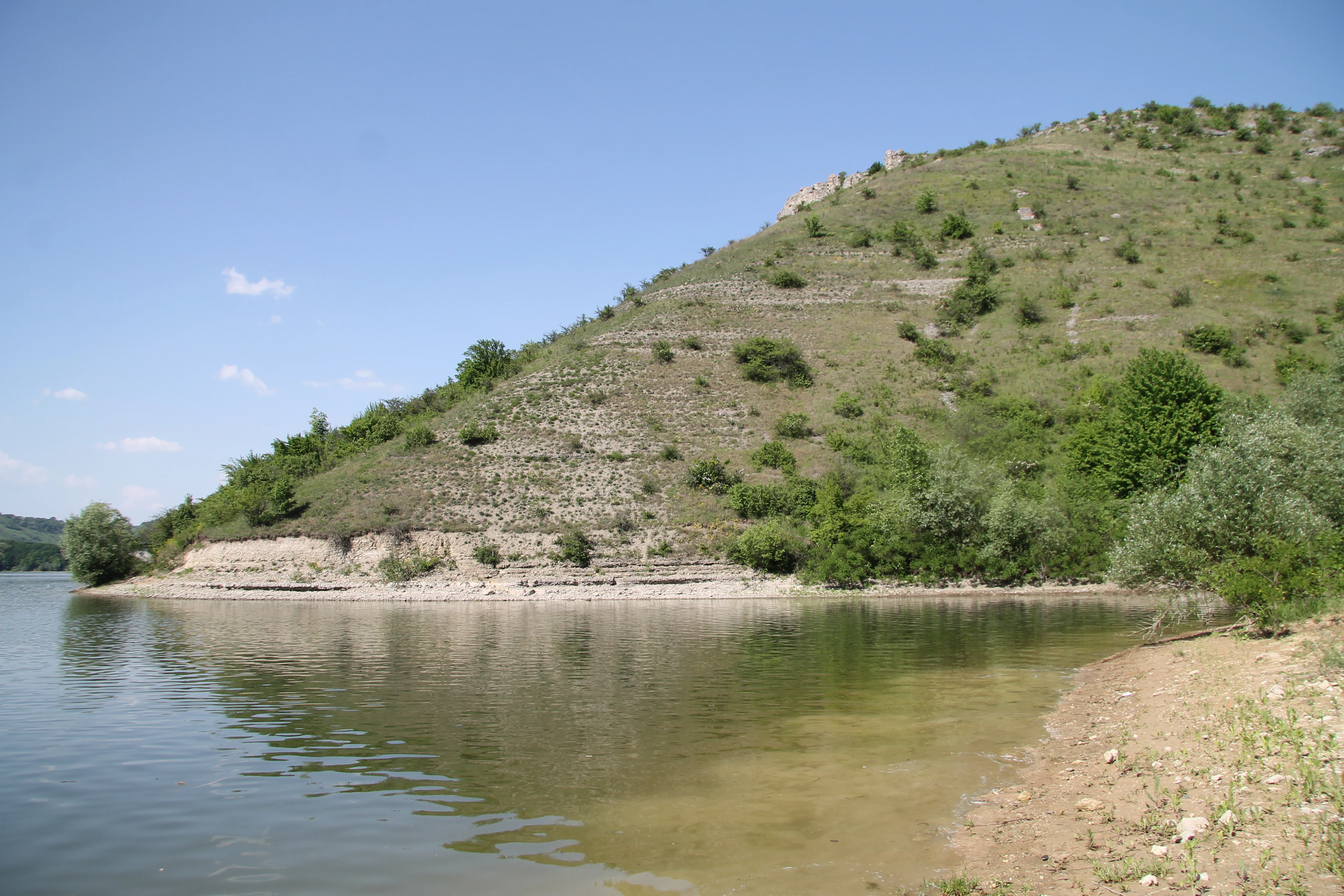 A riverbank with a sloping hillside covered in sparse vegetation and exposed rock layers reflecting in calm water under a clear blue sky